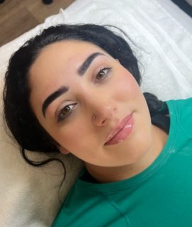Close-up of a young woman with dark hair, lying on a white pillow, wearing a green top, smiling softly at the camera.