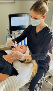 A healthcare professional in black scrubs and a face mask administering a treatment to a patient lying in a hospital bed.
