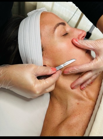 A woman receiving a facial treatment, lying down with a white headband, as a skincare professional uses a tool on her face.