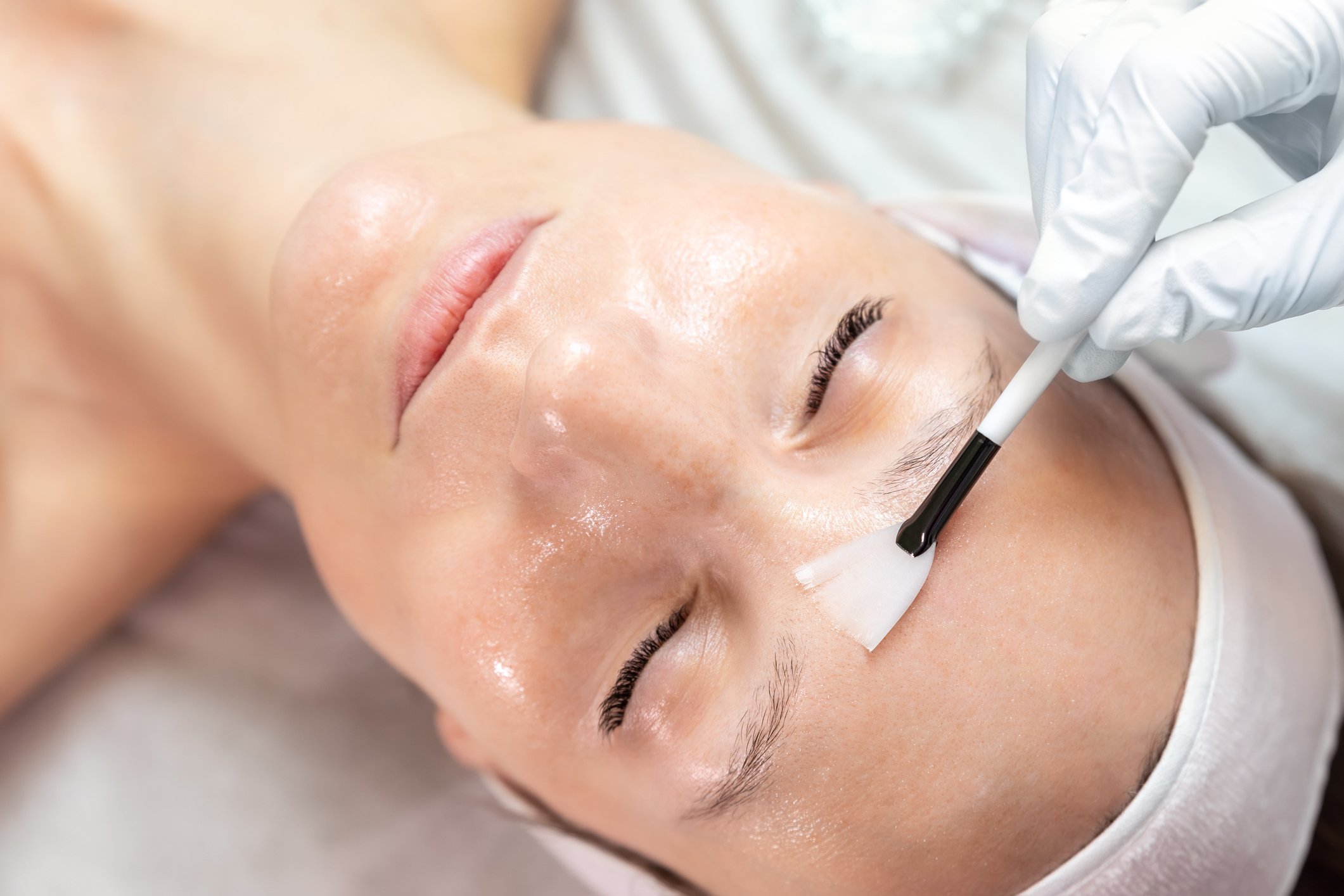 Close-up of a person receiving a facial treatment, with a brush applying a liquid to their forehead, wearing a headband and gloves.