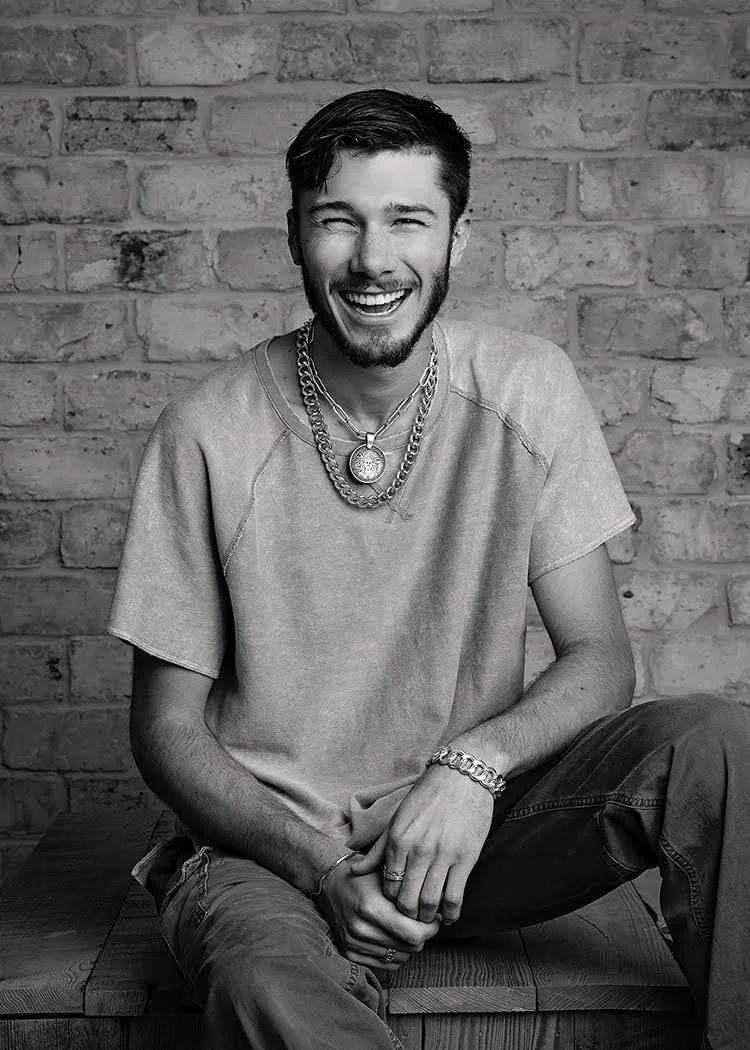 A young man with dark hair and a beard sitting on a bench in front of a brick wall, smiling and laughing, wearing a light t-shirt and layered necklaces.