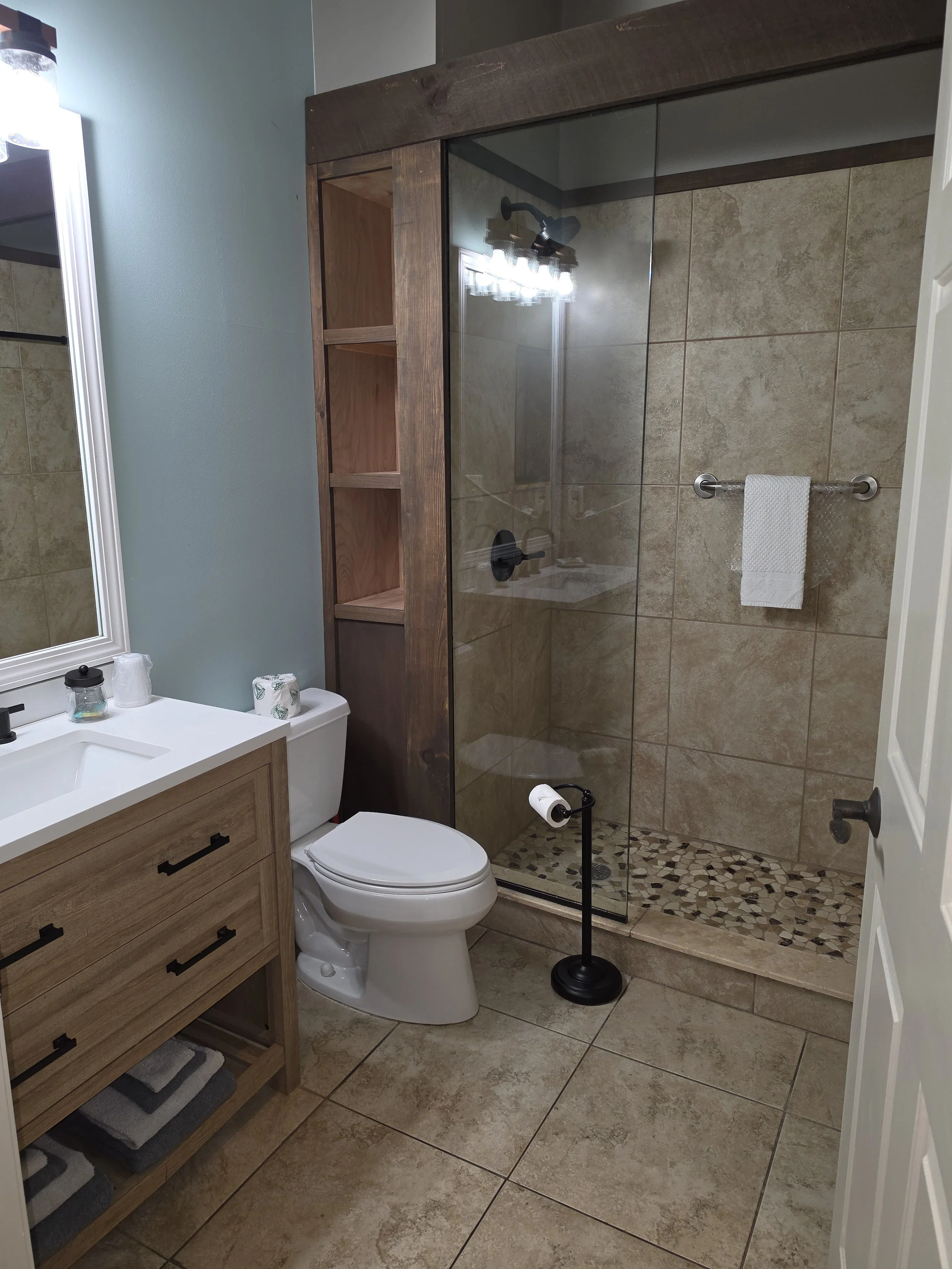 Bathroom with a wooden vanity, a toilet, a glass shower with beige tiles and pebble flooring, a mirror, and a wall-mounted light fixture.