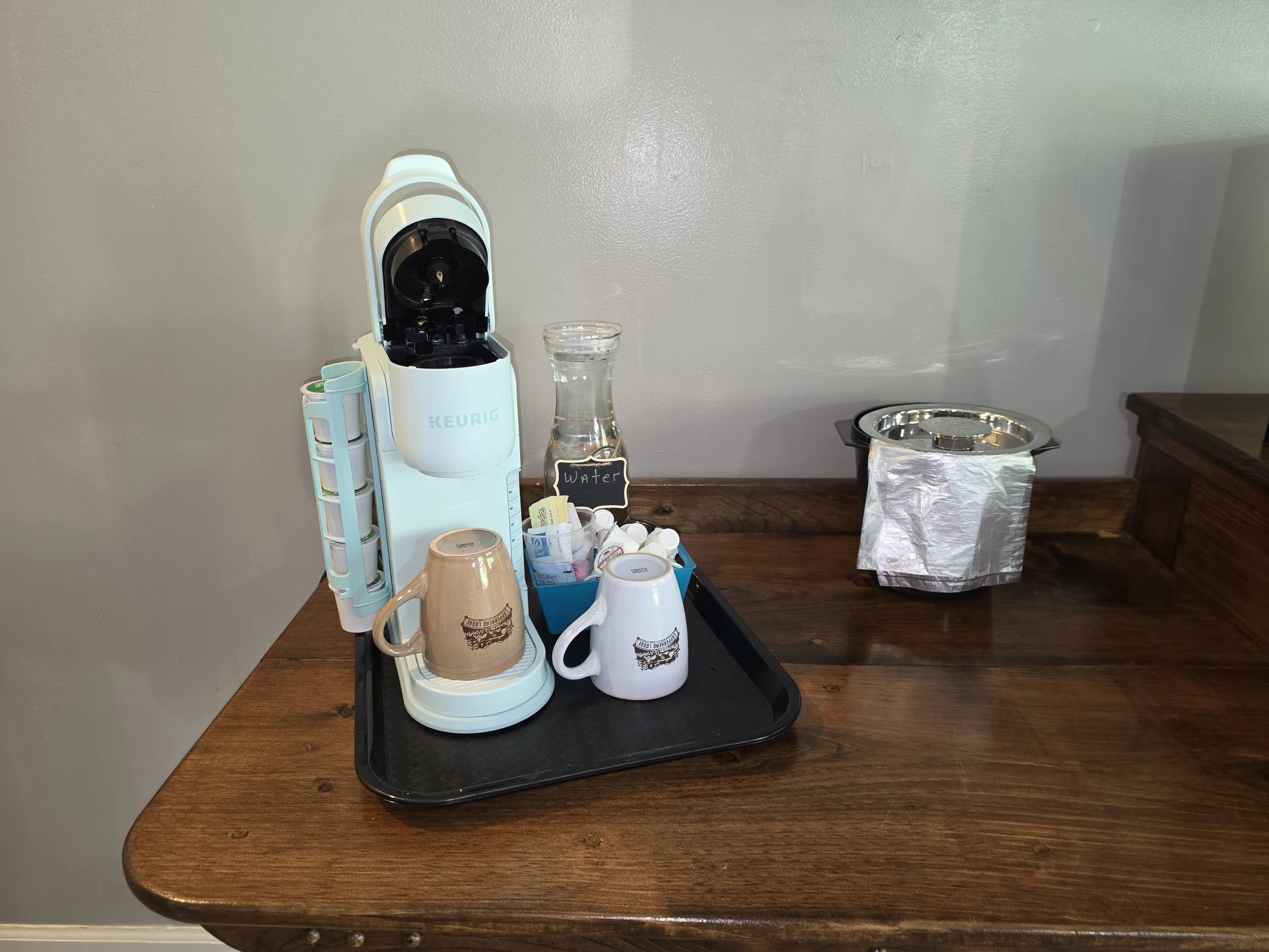A Keurig coffee machine with a glass water vessel, mugs, and various coffee supplies on a black tray on a wooden table against a light-colored wall.