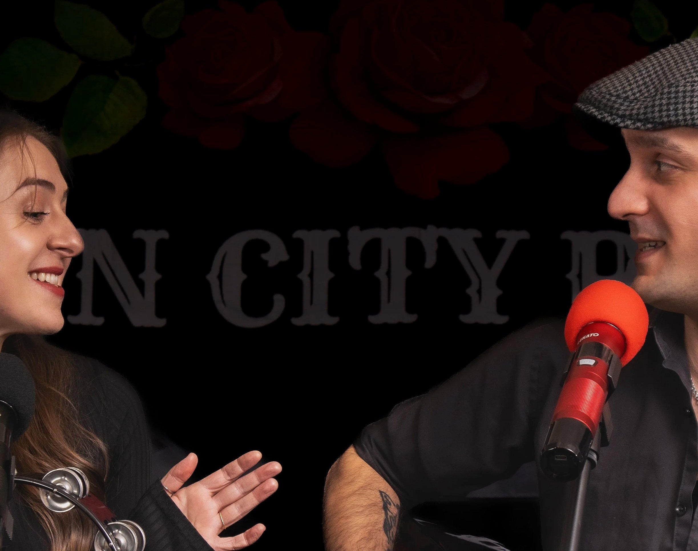 A woman and a man in a radio studio having a conversation, with a dark background that reads "IN CITY" and red and black microphones in front of them.
