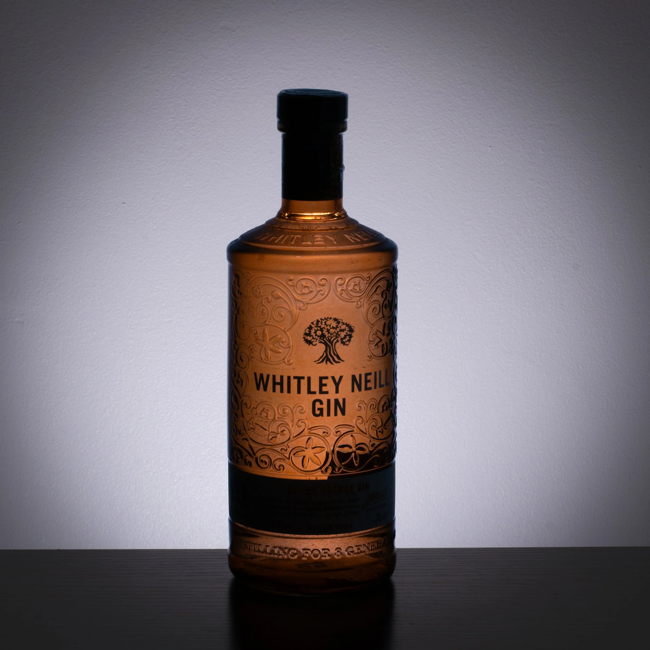 Backlit bottle of Whitley Neill gin on a dark surface with a neutral background.