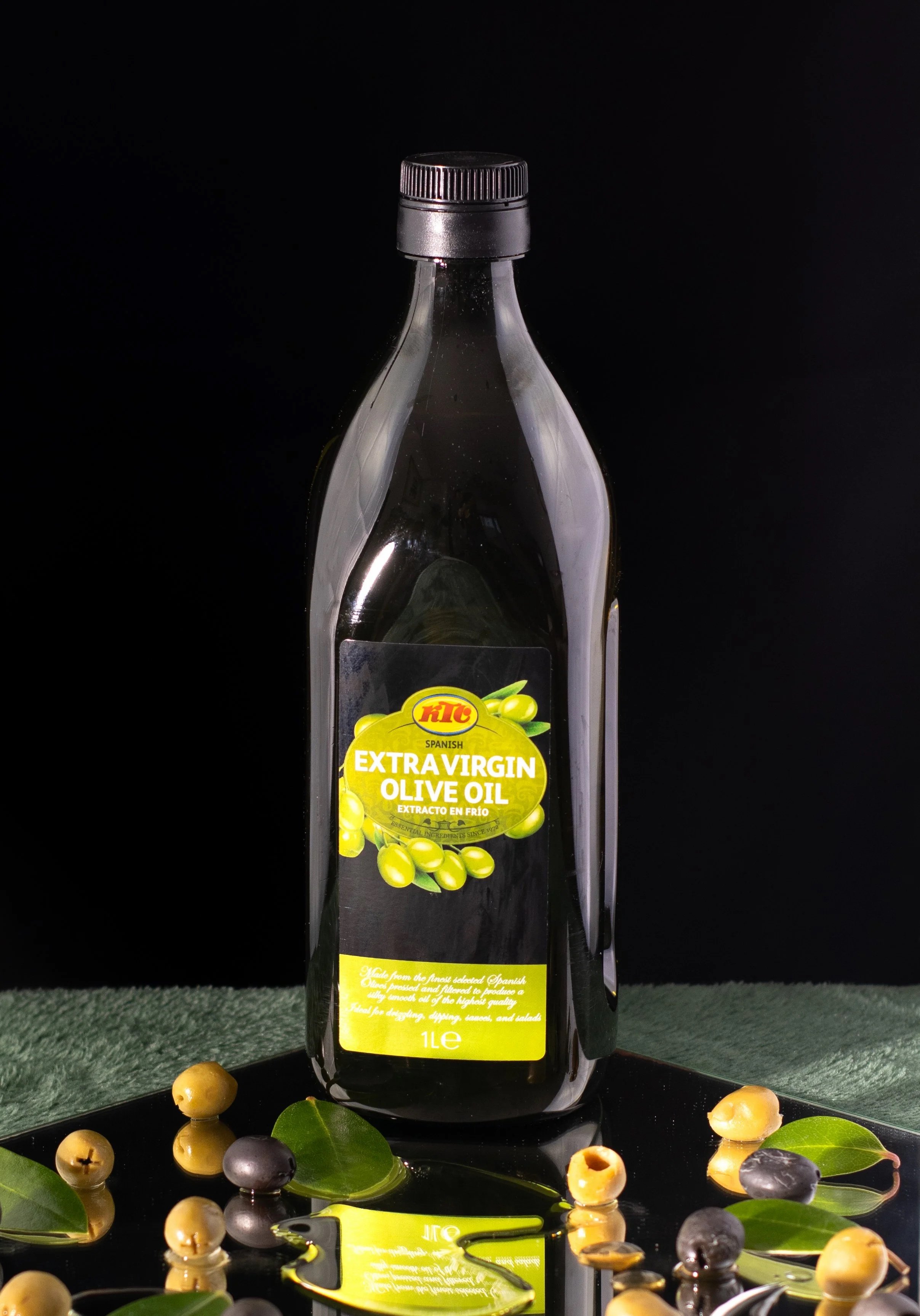 A 1-liter bottle of Spanish extra virgin olive oil with a black cap, placed on a reflective black surface surrounded by green leaves and mixed green and black olives against a black background.
