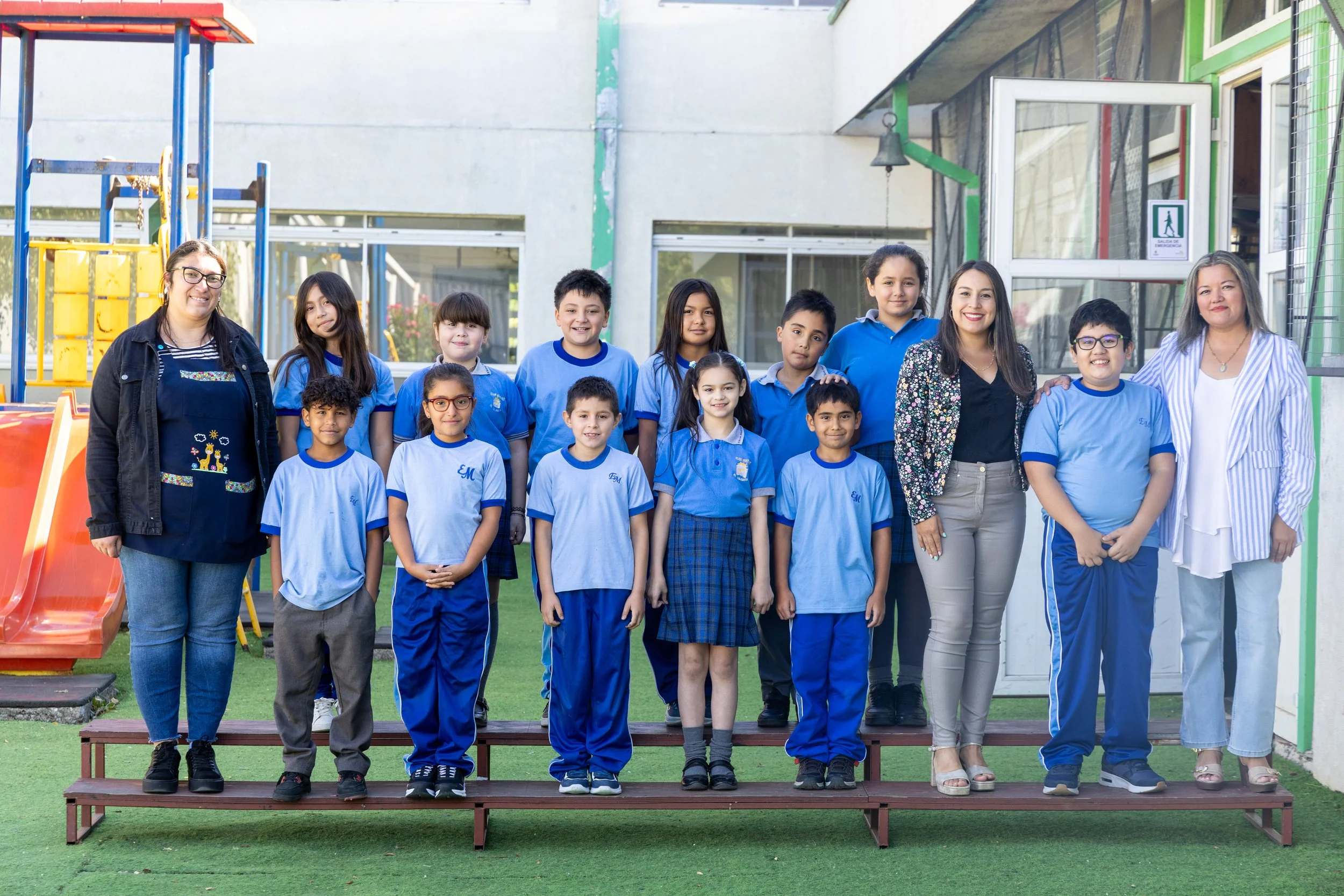 Grupo de niños y adultos en una escuela, personas de pie en una fila en un patio escolar con equipo de juego y edificios en el fondo.