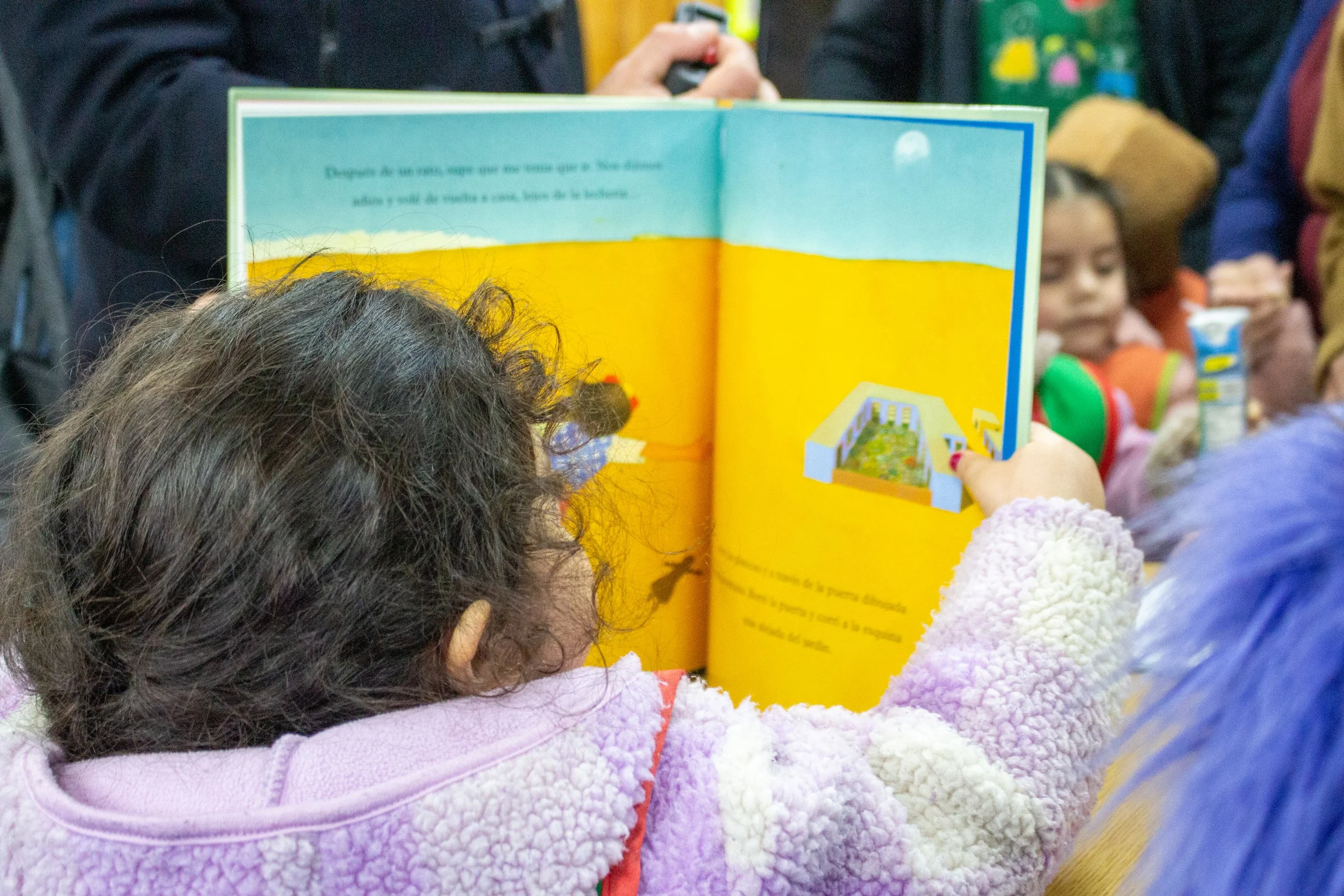 Niña leyendo un libro con ilustraciones a un grupo de niños en un entorno escolar o de biblioteca.
