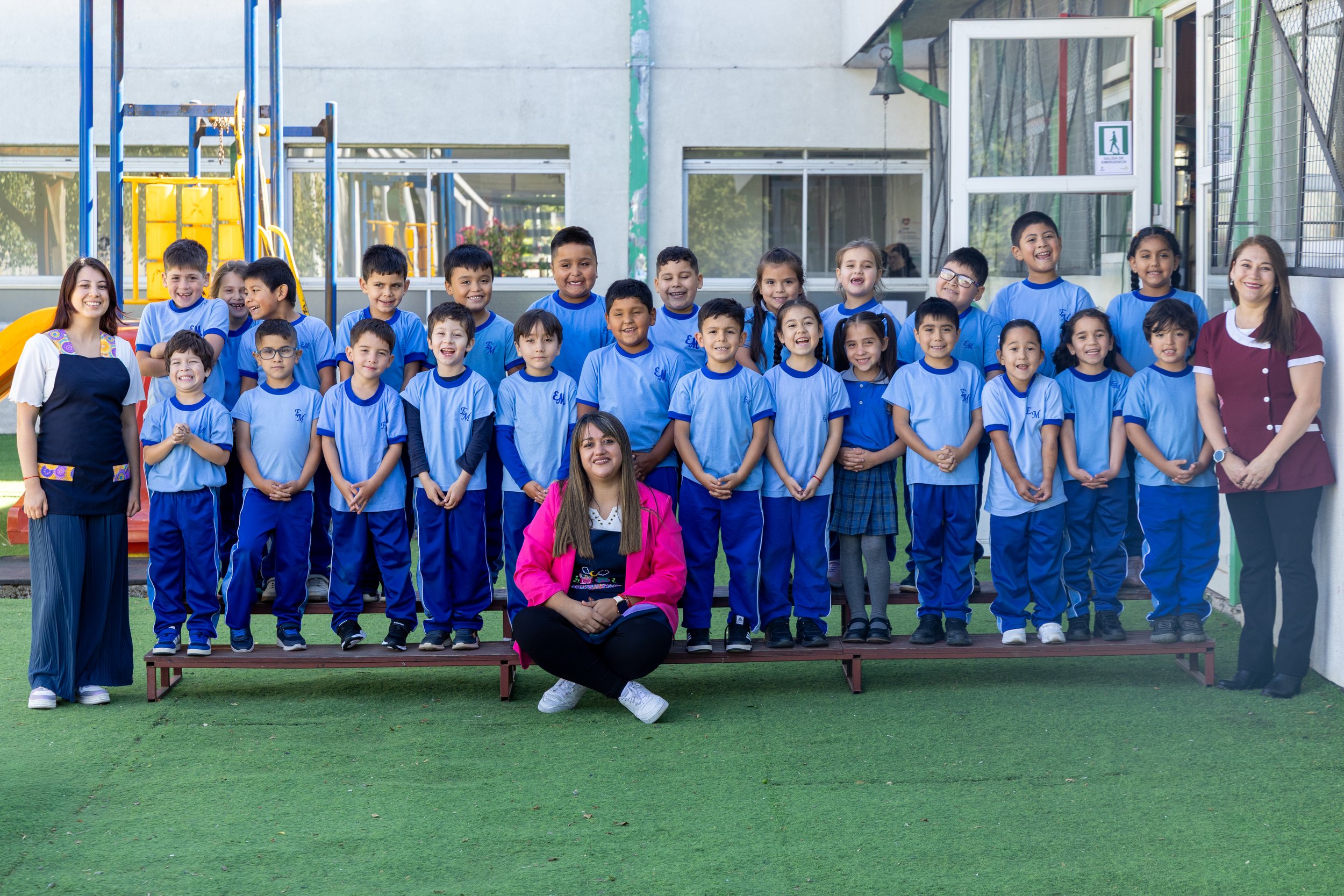 Grupo de niños y niñas con sus maestros en un patio escolar, vestimenta escolar azul, jóvenes sonriendo delante de un patio con juegos y un edificio escolar en el fondo.