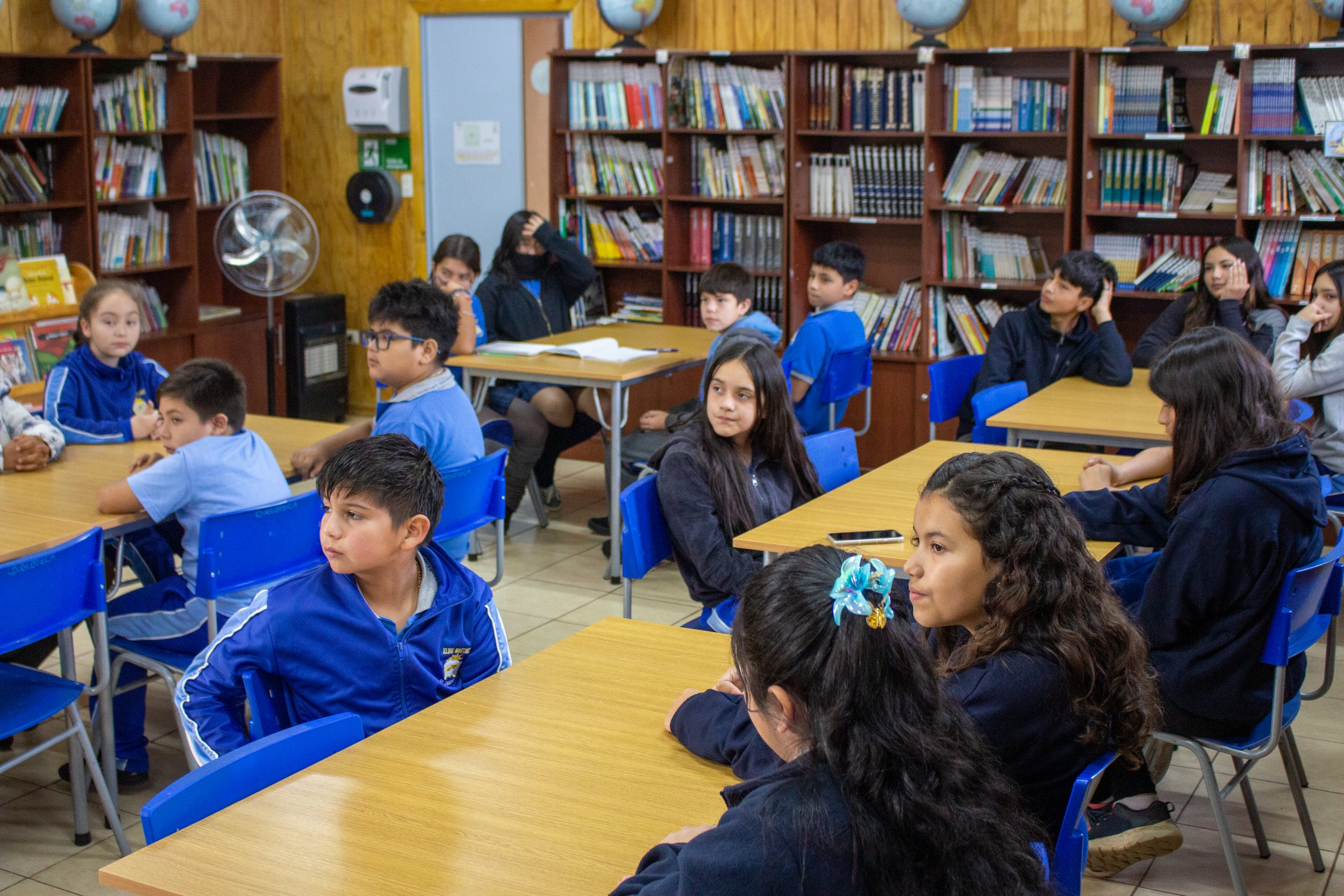 Niños en un aula de biblioteca, sentados en mesas de color madera y sillas azules, rodeados de estanterías llenas de libros, algunos con expresiones de atención, otros distraídos o pensando.
