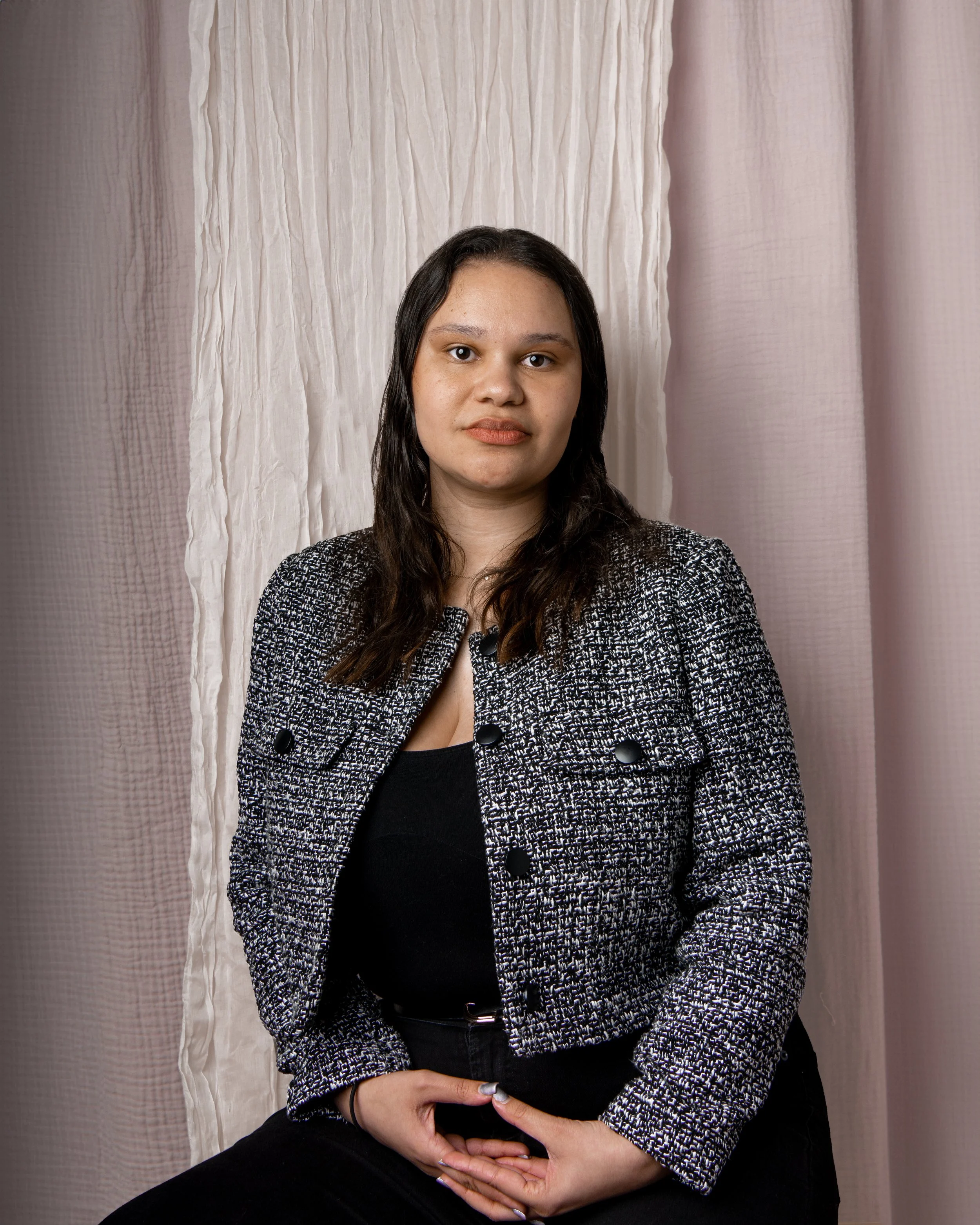 Brunette woman with a grey blazer on sitting with her hands on her lap. The background is pink and white fabric that is textured.