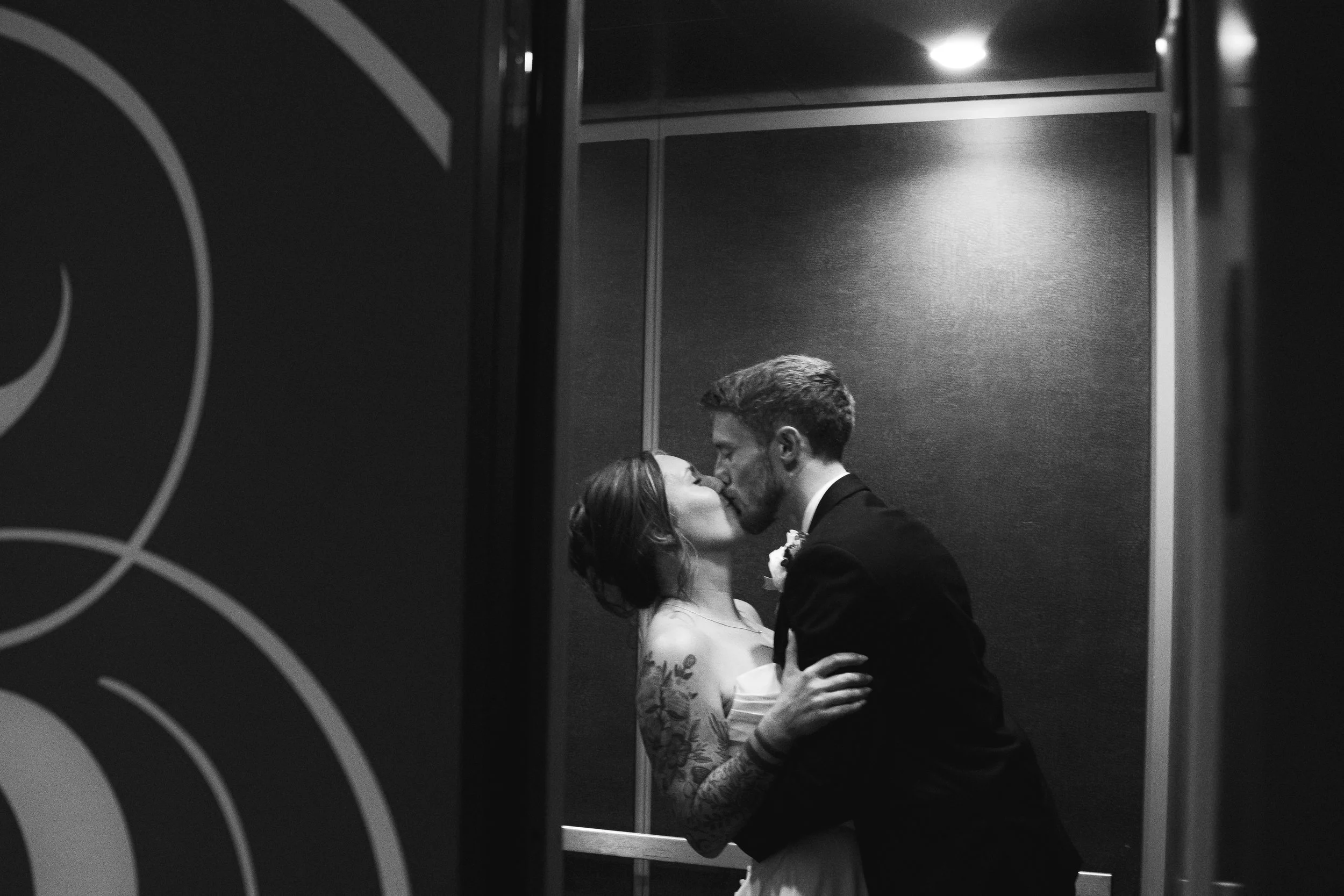 A black-and-white photo of a man and woman kissing in an elevator. The man is dressed in a suit, and the woman is wearing a sleeveless dress with floral tattoos on her arm. Their faces are close together, and the elevator walls are plain with a textu