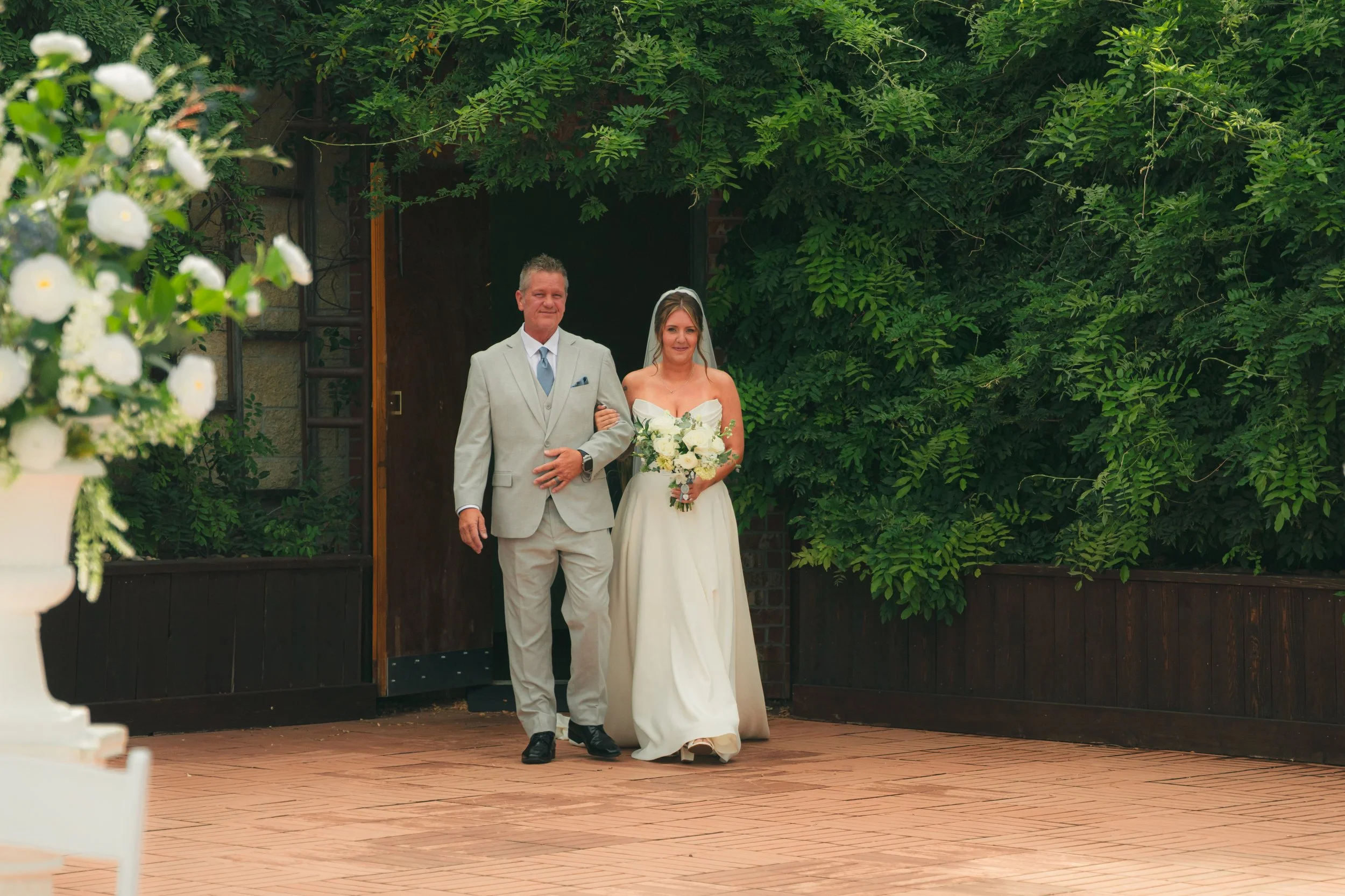 A bride and an older man, possibly her father, walking down the aisle outside during a wedding ceremony. The woman is wearing a white strapless wedding dress and holding a bouquet of white flowers. The man is dressed in a light-colored suit with a ti