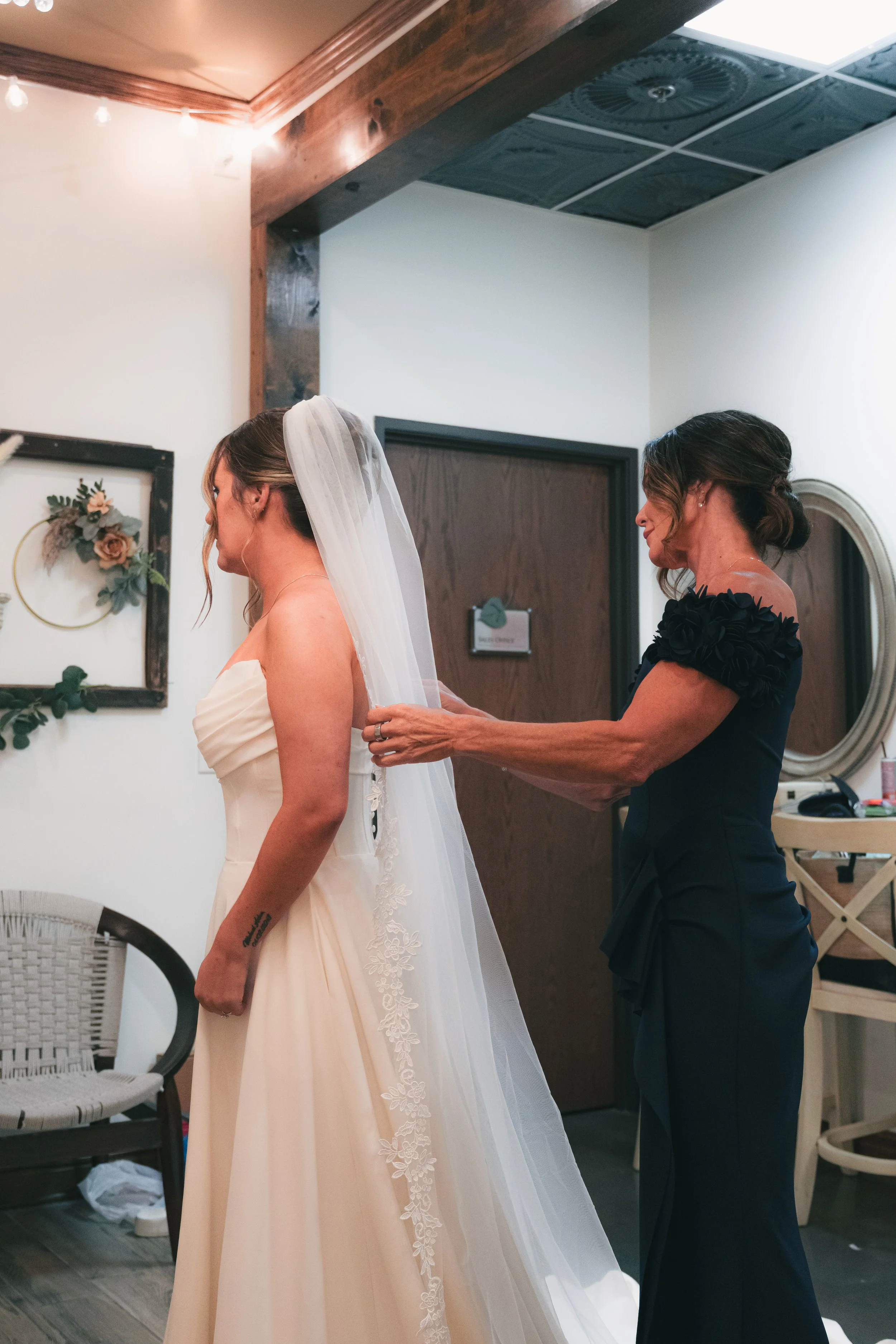 A bride in a light-colored wedding dress with a veil being helped by a woman in a dark dress, in an indoor setting decorated with floral art and string lights.