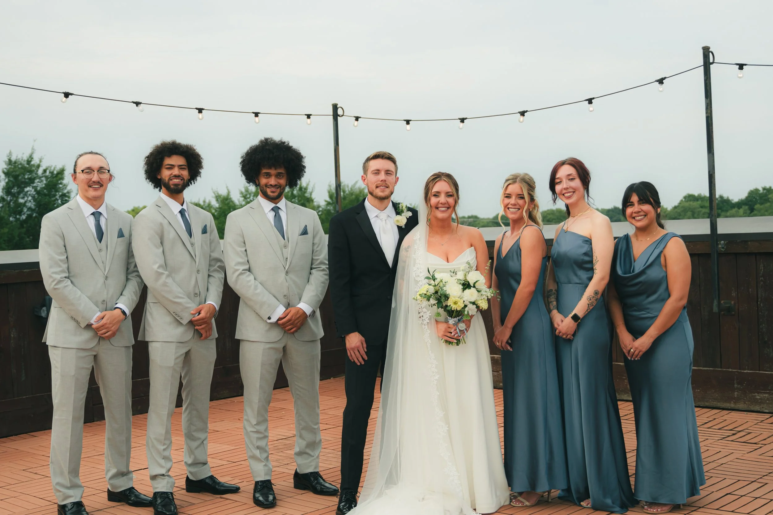 Group of people dressed in wedding attire standing outdoors on a rooftop with string lights, smiling at the camera.