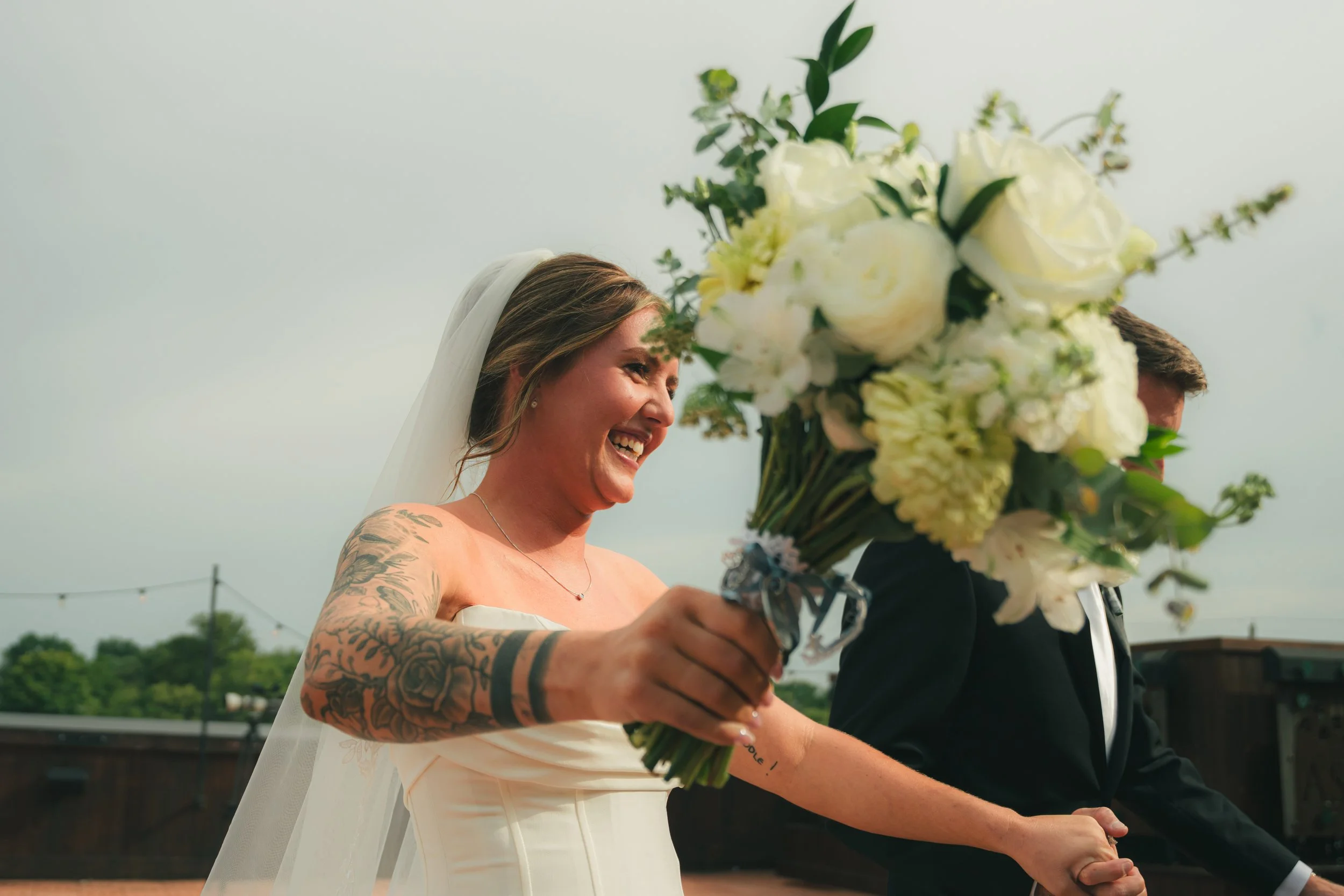 A bride with tattoos smiling and holding a large bouquet of white flowers during a wedding ceremony, with a light blue sky and greenery in the background.