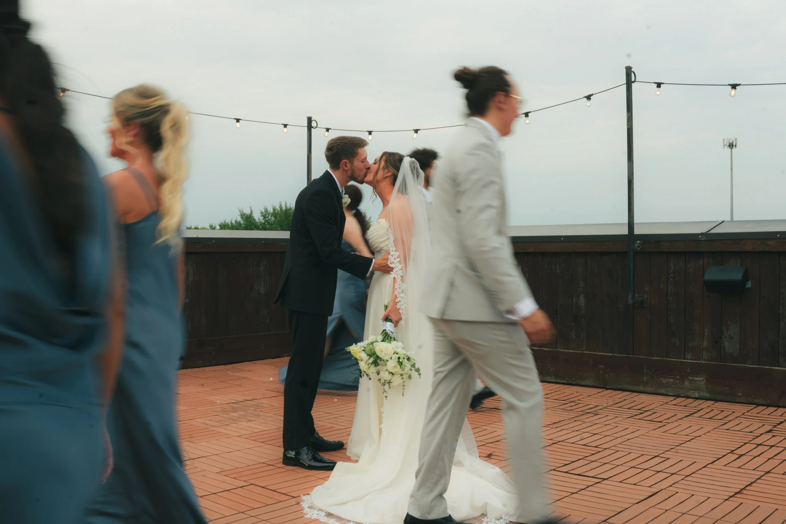 A wedding reception taking place outdoors with string lights overhead. A couple is kissing, the bride in a white wedding dress and veil, holding a bouquet of flowers, and the groom in a dark tuxedo. Several guests are walking in the foreground, some 
