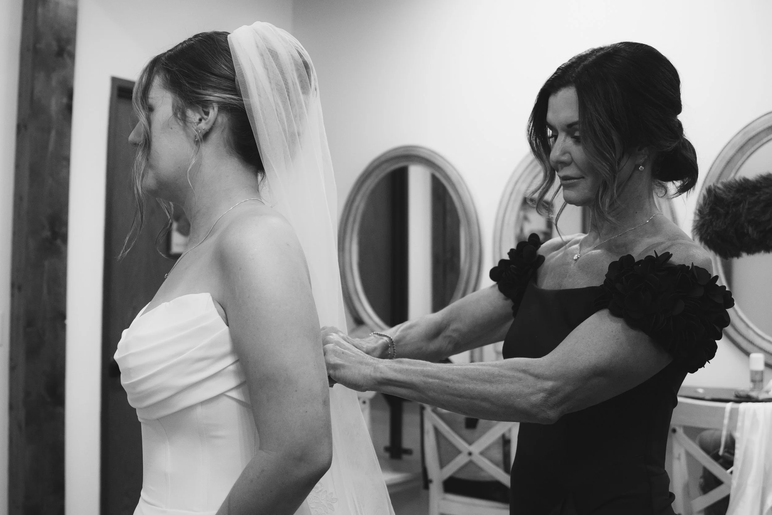 A bride in a white wedding dress and veil is being assisted by a woman in a black dress, possibly her mother or a stylist, as she prepares for the wedding. The scene is in a room with mirrors and furniture.