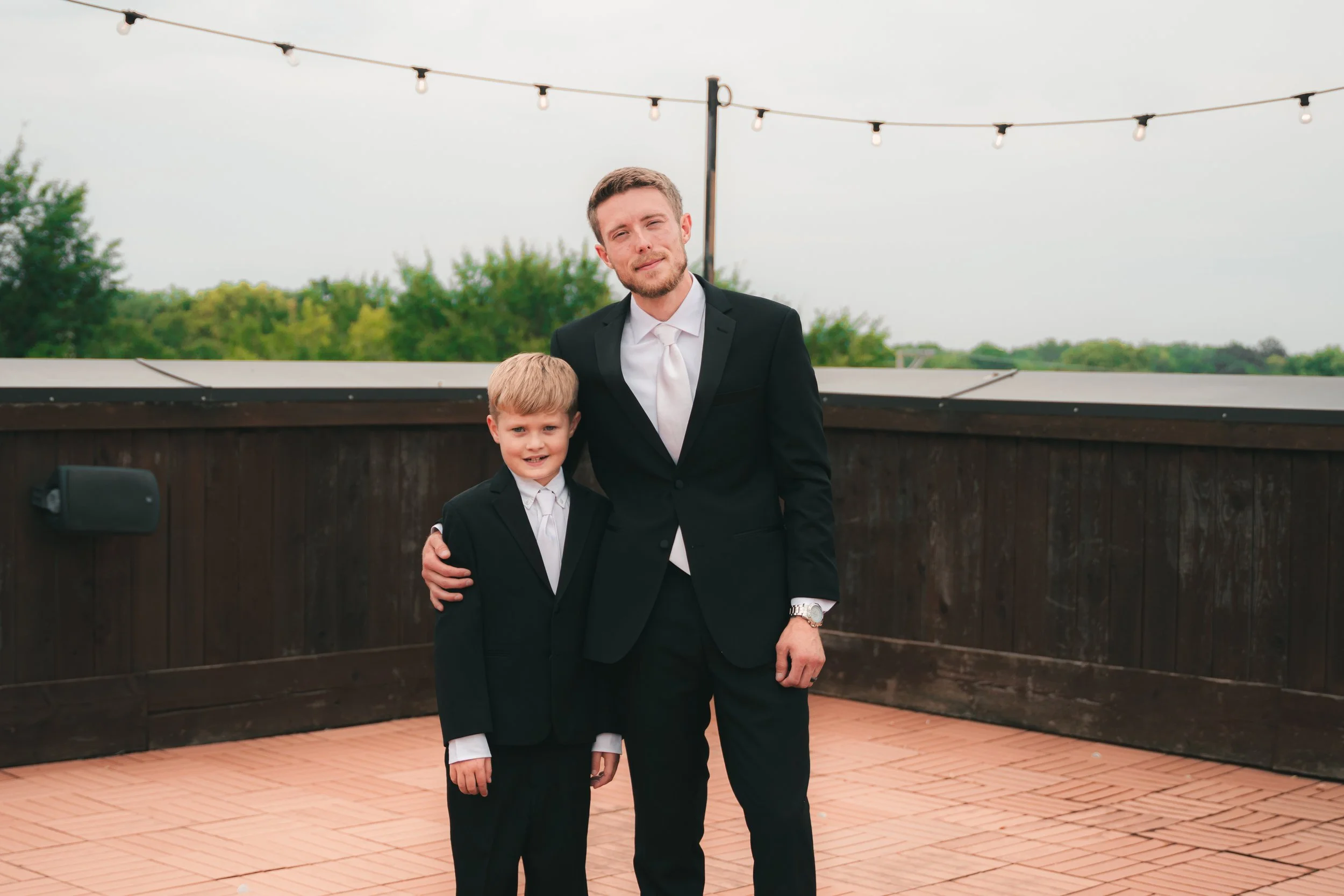 A man and a young boy dressed in formal suits standing together outdoors on a patio with string lights overhead. The man has a beard and short hair, and the boy has blond hair. Trees are visible in the background.
