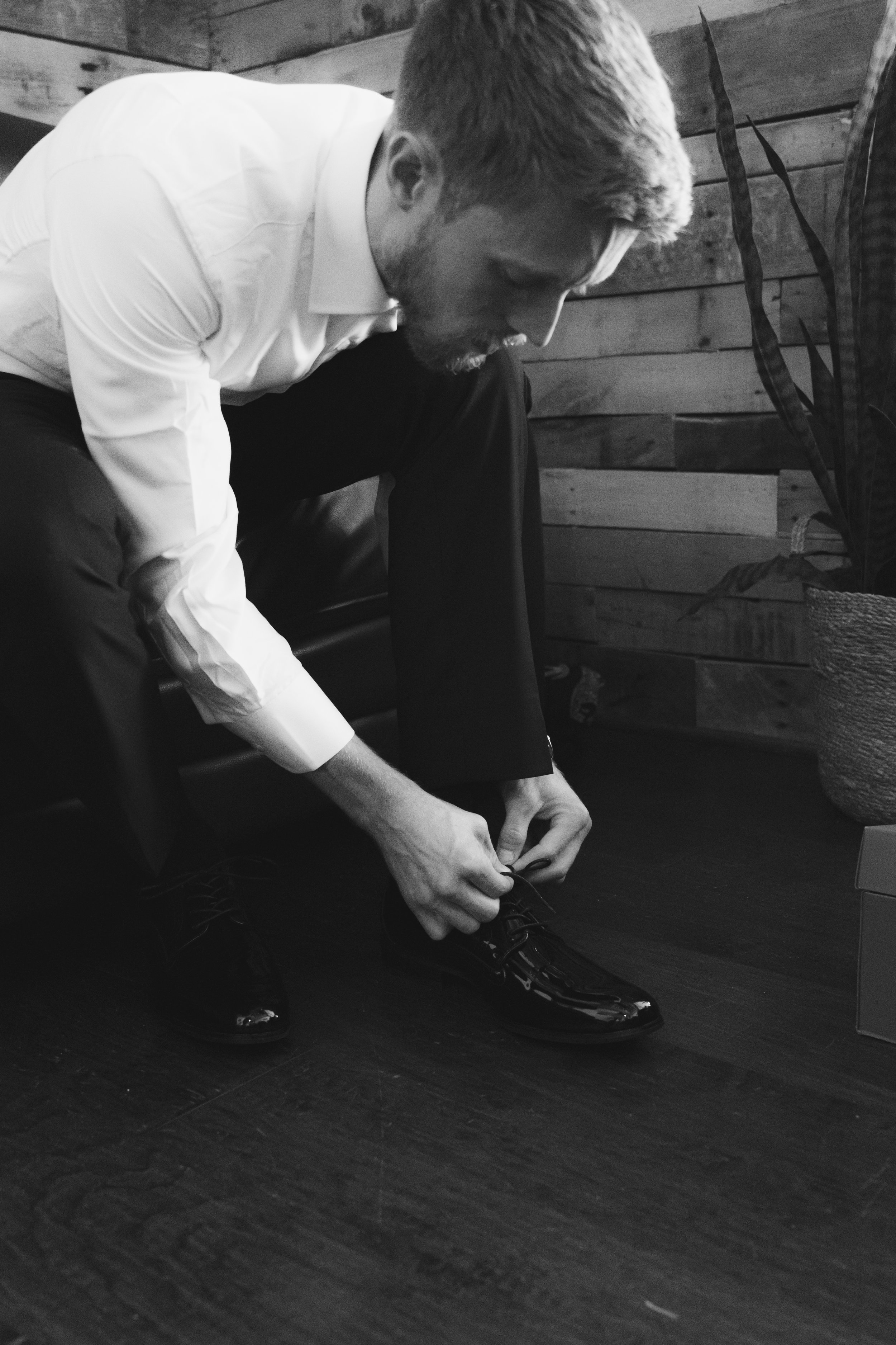 A man sitting on a chair, tying his shoelaces, in a room with wooden walls and a plant in a basket.