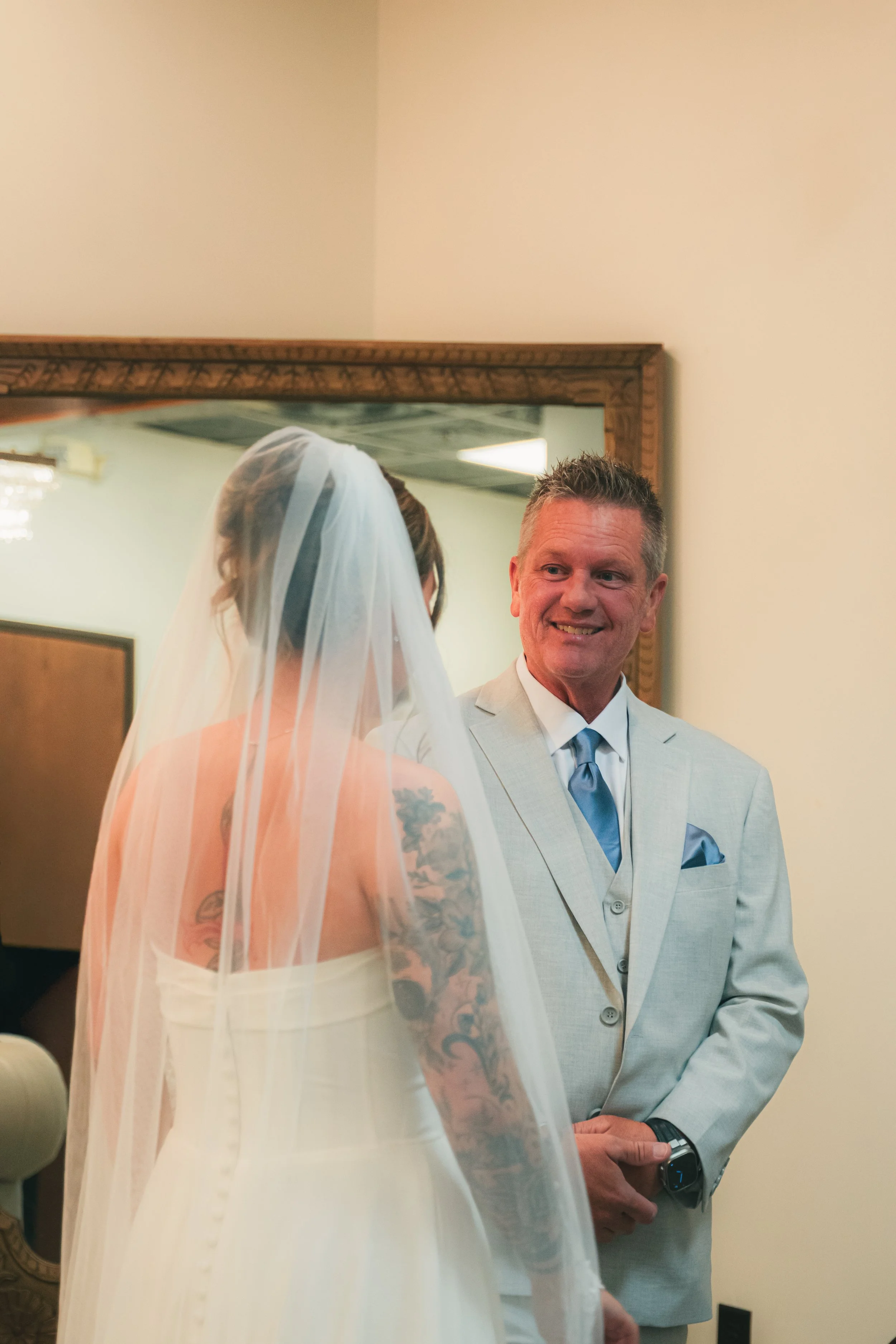 A bride in a white wedding dress with tattoos on her back and arms and a sheer veil is facing a man in a light-colored suit with a blue tie, who is smiling.