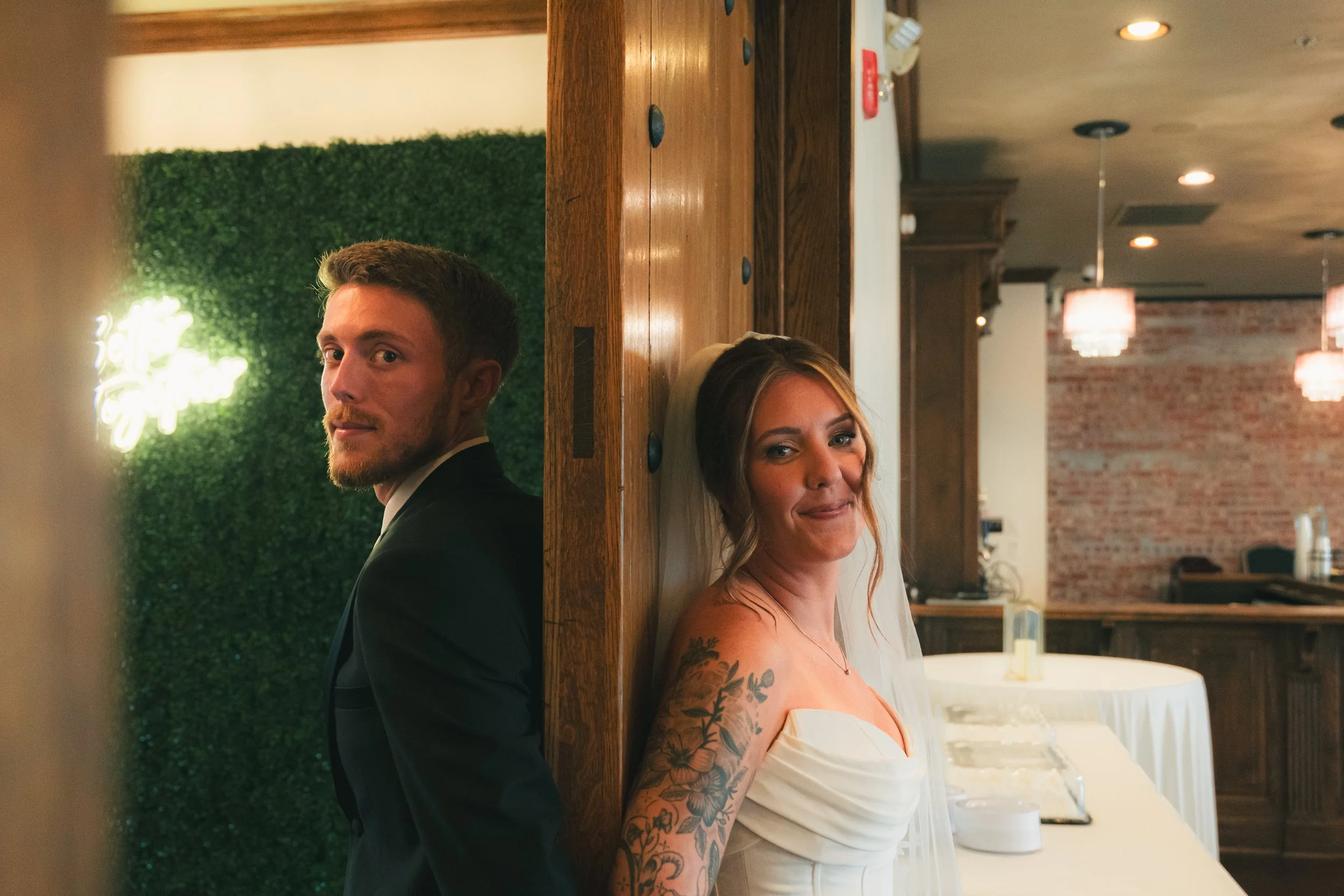 A young man in a suit and a young woman in a strapless dress are standing back-to-back, separated by a wooden divider, in a decorated interior space with green wall and brick wall backgrounds.