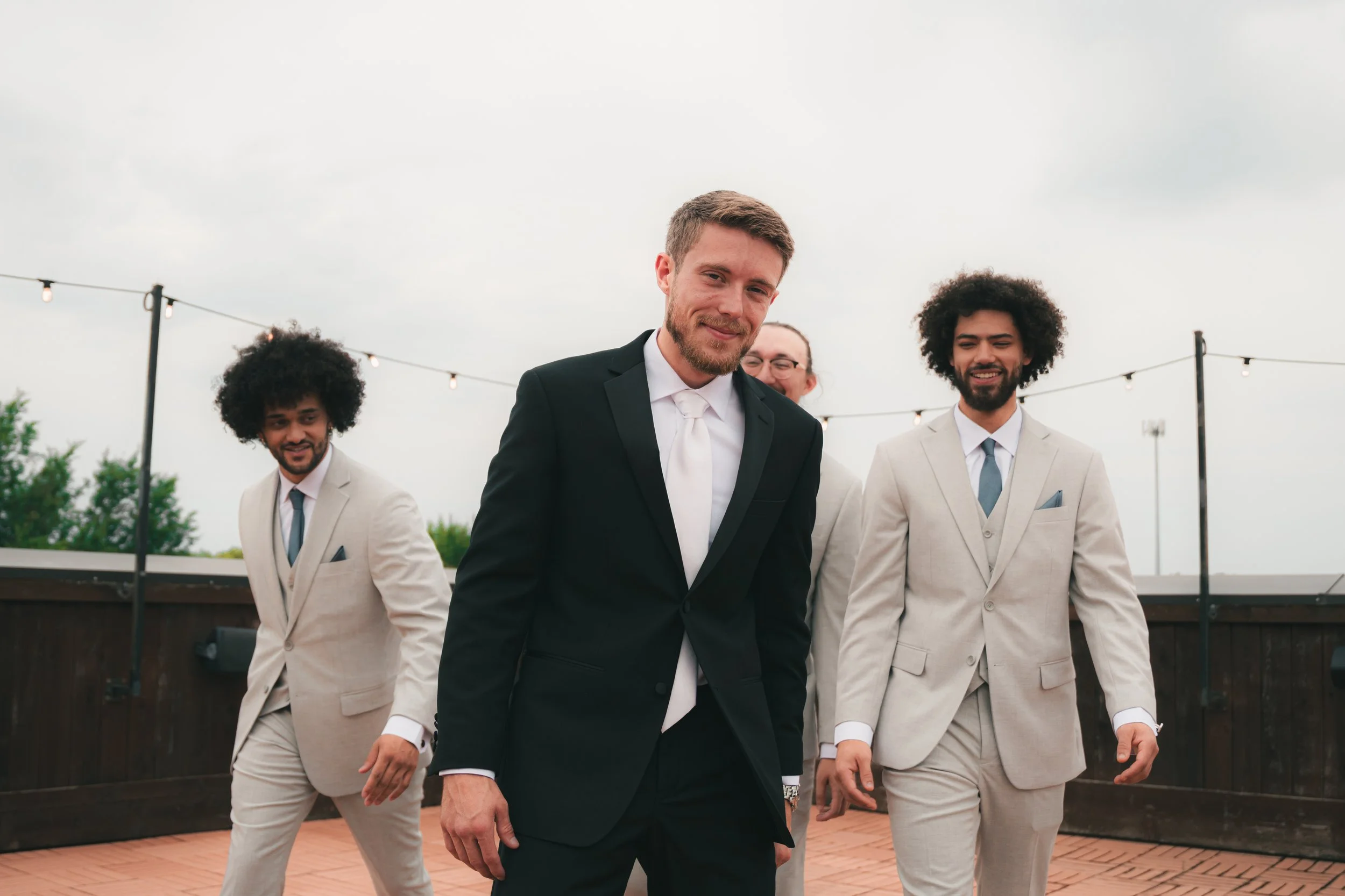 Four men dressed in formal suits walking outdoors on a patio with string lights overhead. The man in front wears a black suit with a white tie, while the others wear light-colored suits.