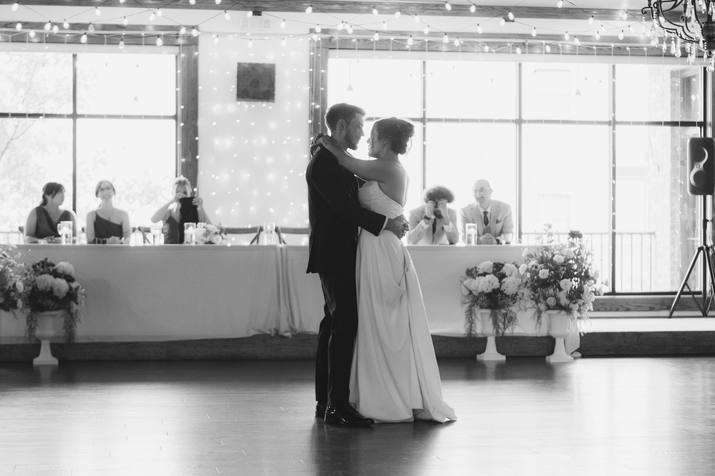A couple shares a dance at their wedding reception, with guests seated at a decorated table in the background and large windows letting in natural light.