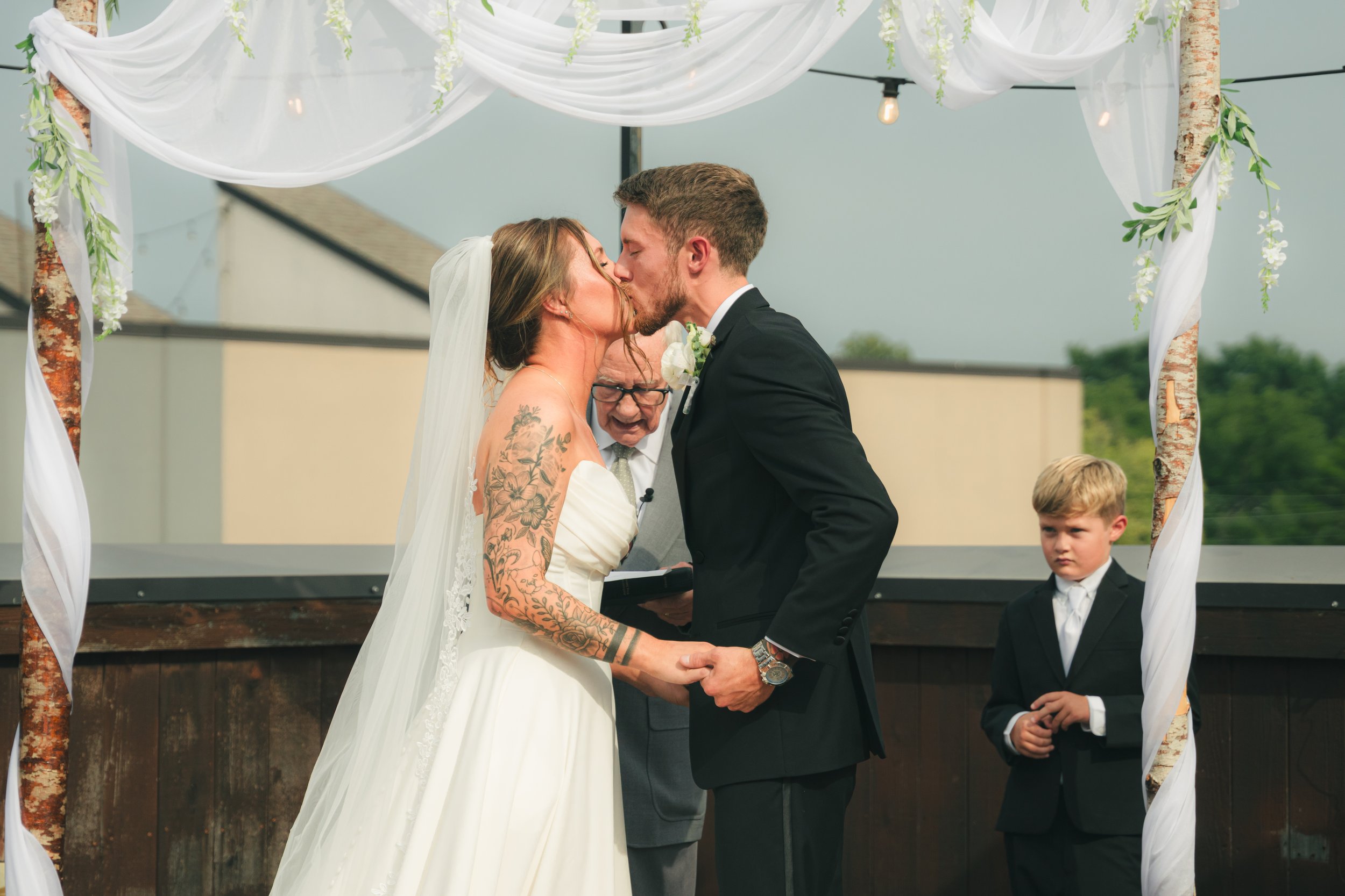 A bride and groom kiss during their outdoor wedding ceremony, with an officiant holding a book and a young boy in a suit standing nearby, under a decorated arch with white drapery and green foliage.