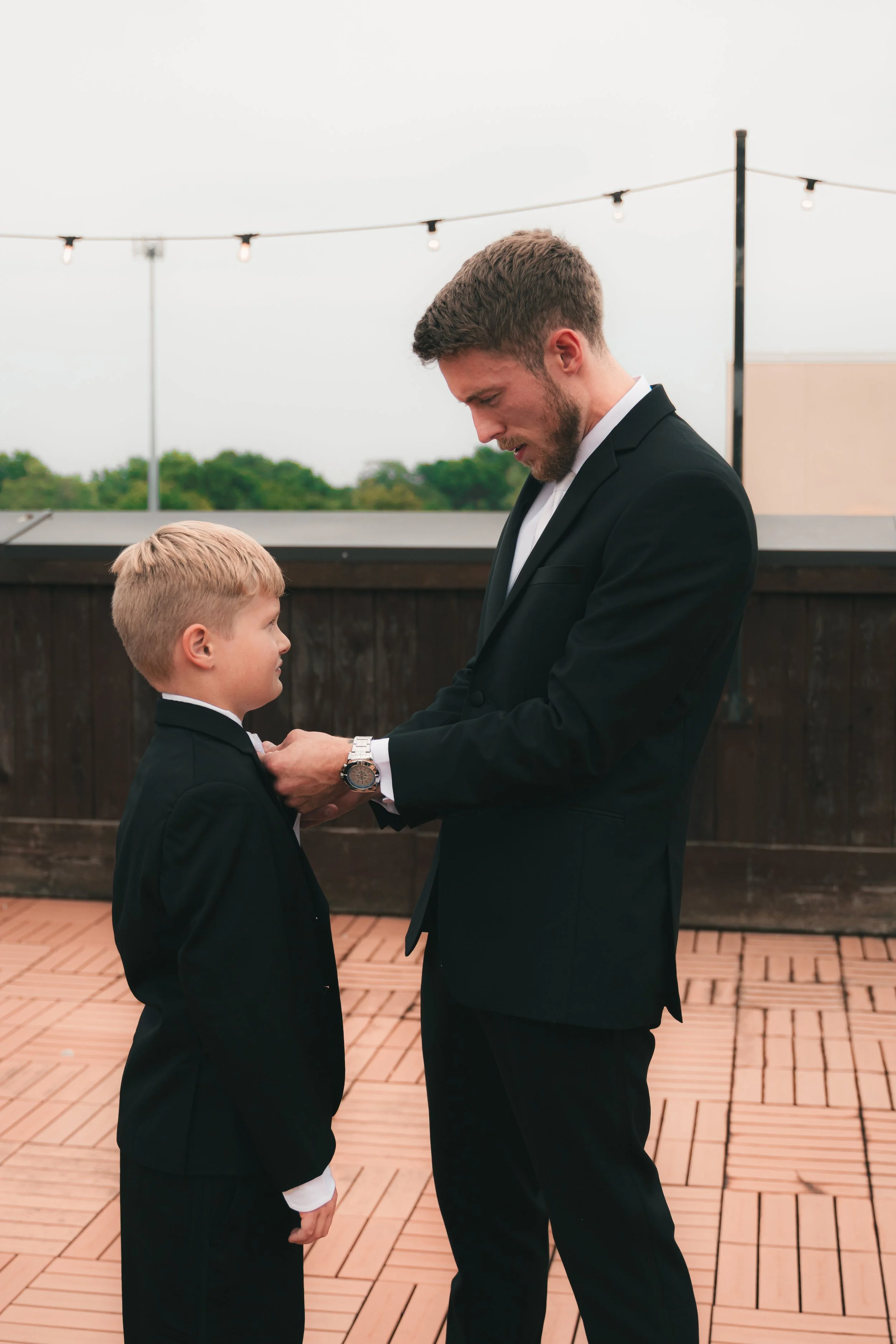 A man and a boy in formal suits holding hands on an outdoor patio, with string lights overhead and green trees in the background.