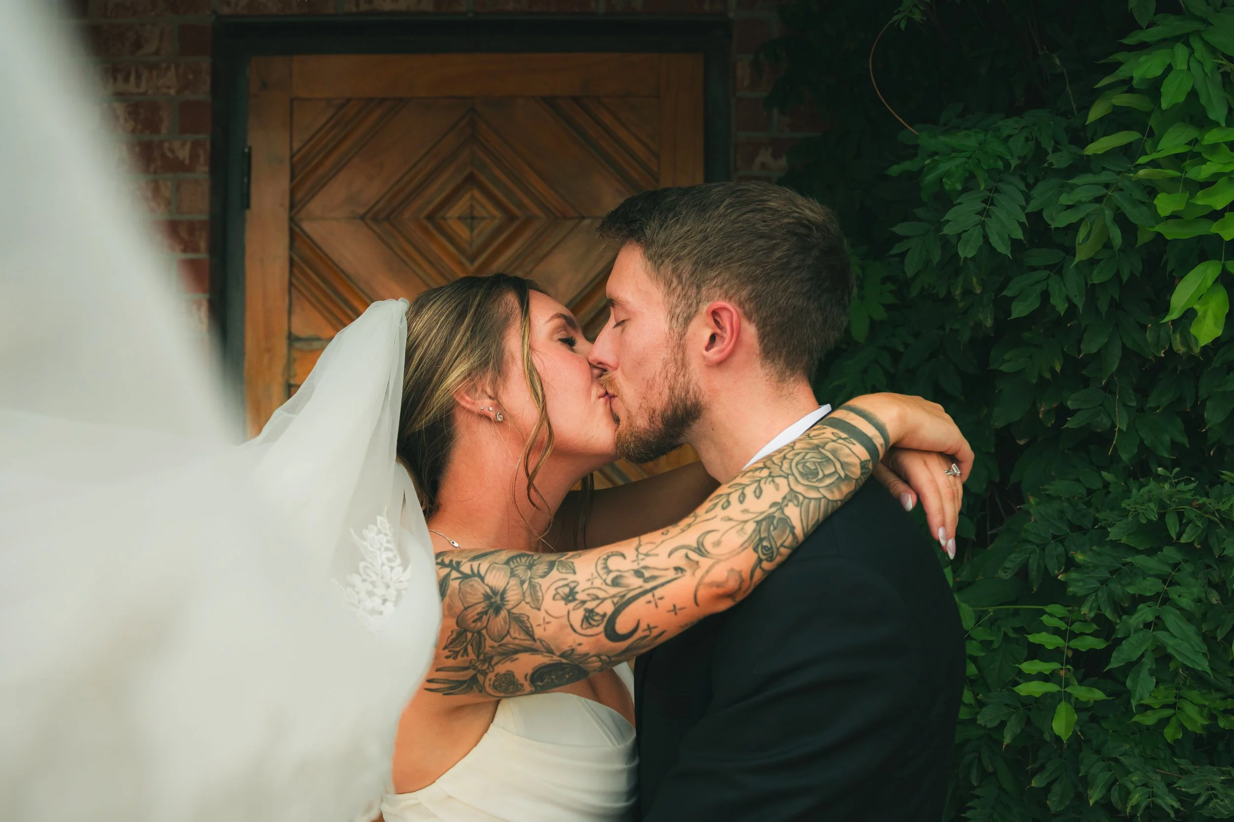A bride and groom kiss during their wedding in front of a wooden door and green foliage.