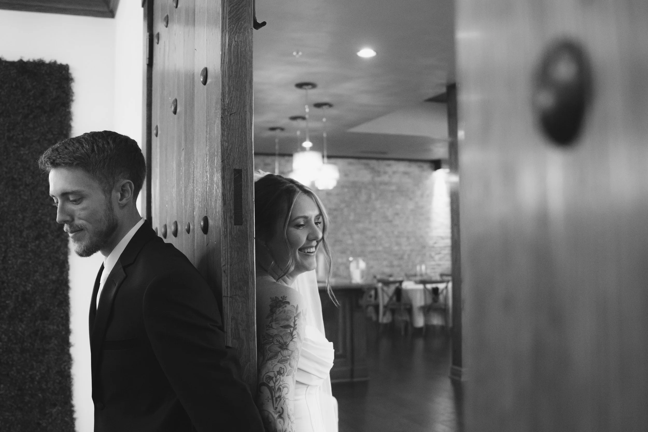 Black and white photo of a man in a suit and a woman in a wedding dress standing shoulder to shoulder behind a wooden partition, smiling and looking down in a warmly lit indoor setting.