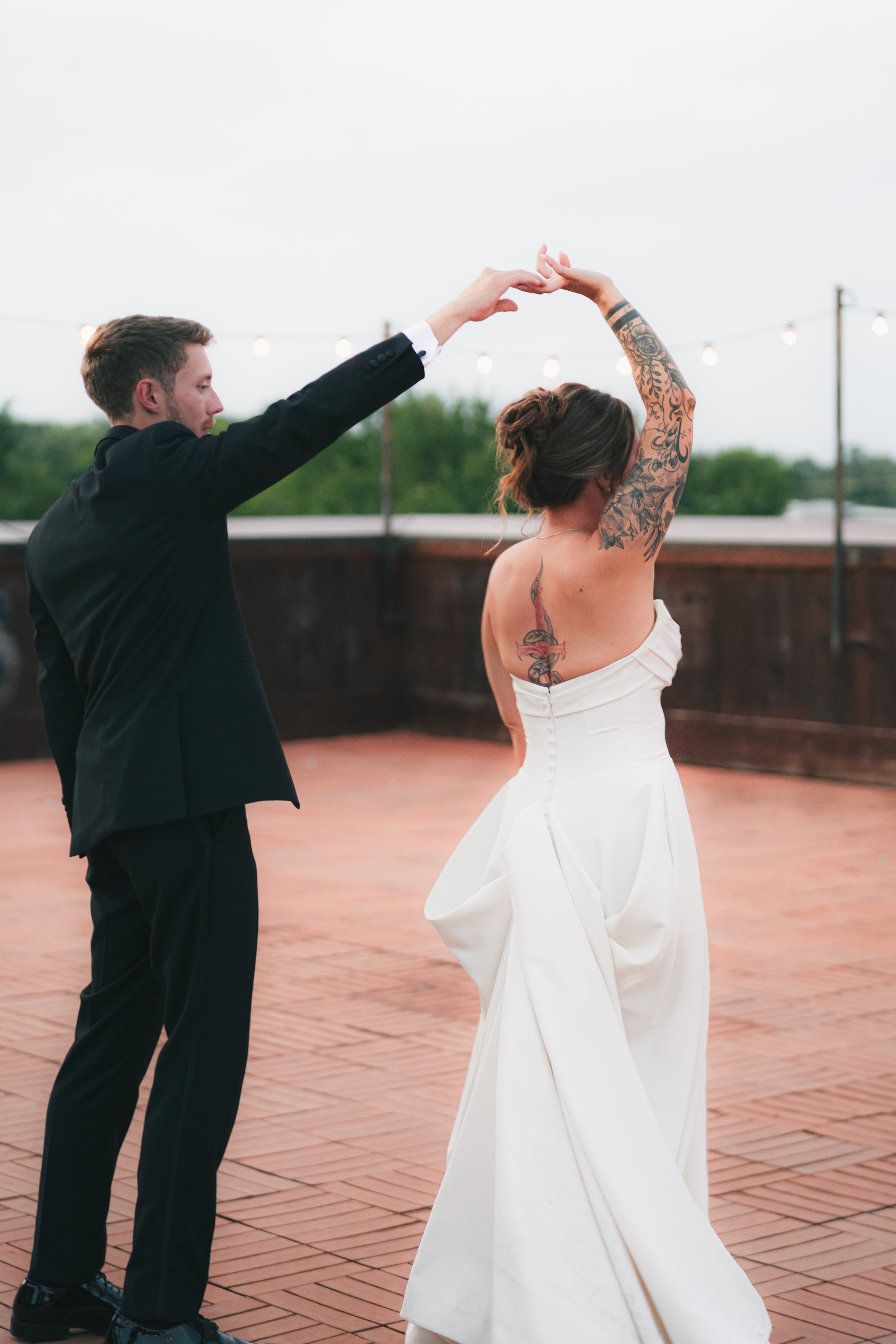 A couple dancing on an outdoor wooden deck during evening, with string lights overhead, the woman in a strapless white dress with tattoos on her arm and back, and the man in a black suit, holding hands and dancing