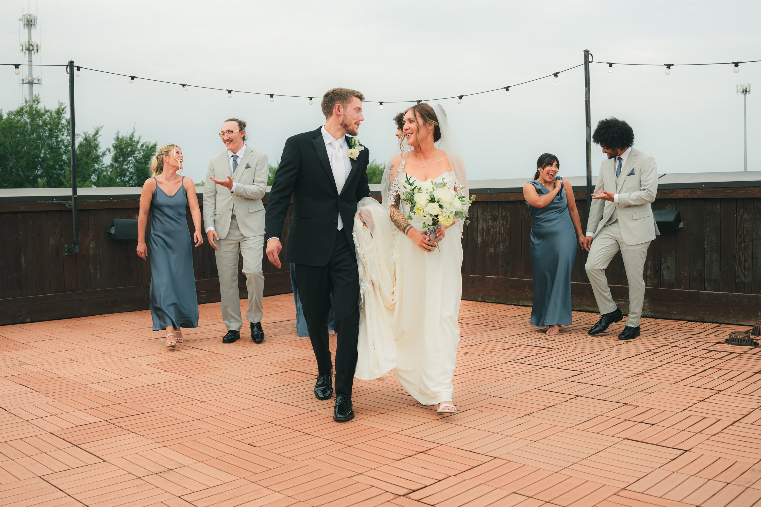 A group of wedding guests dancing on an outdoor terrace with string lights, including a bride and groom in the center, with other women in blue dresses and men in light gray suits.