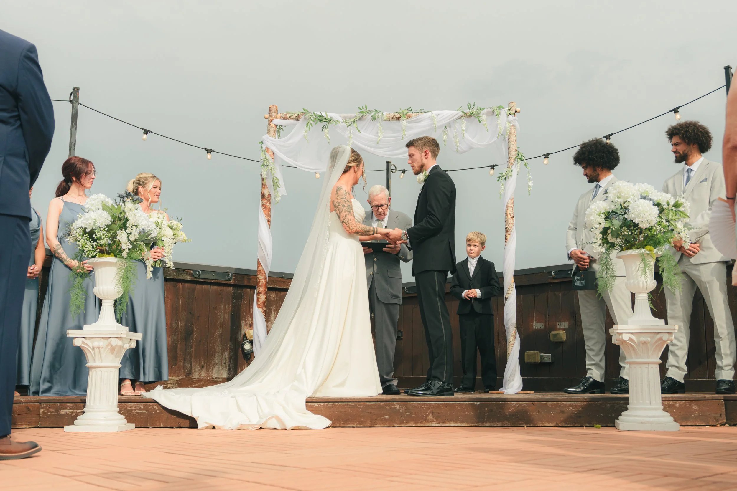 A couple gets married on an outdoor ceremony stage decorated with white fabric and greenery, surrounded by their wedding party, on a wooden deck, with string lights overhead.