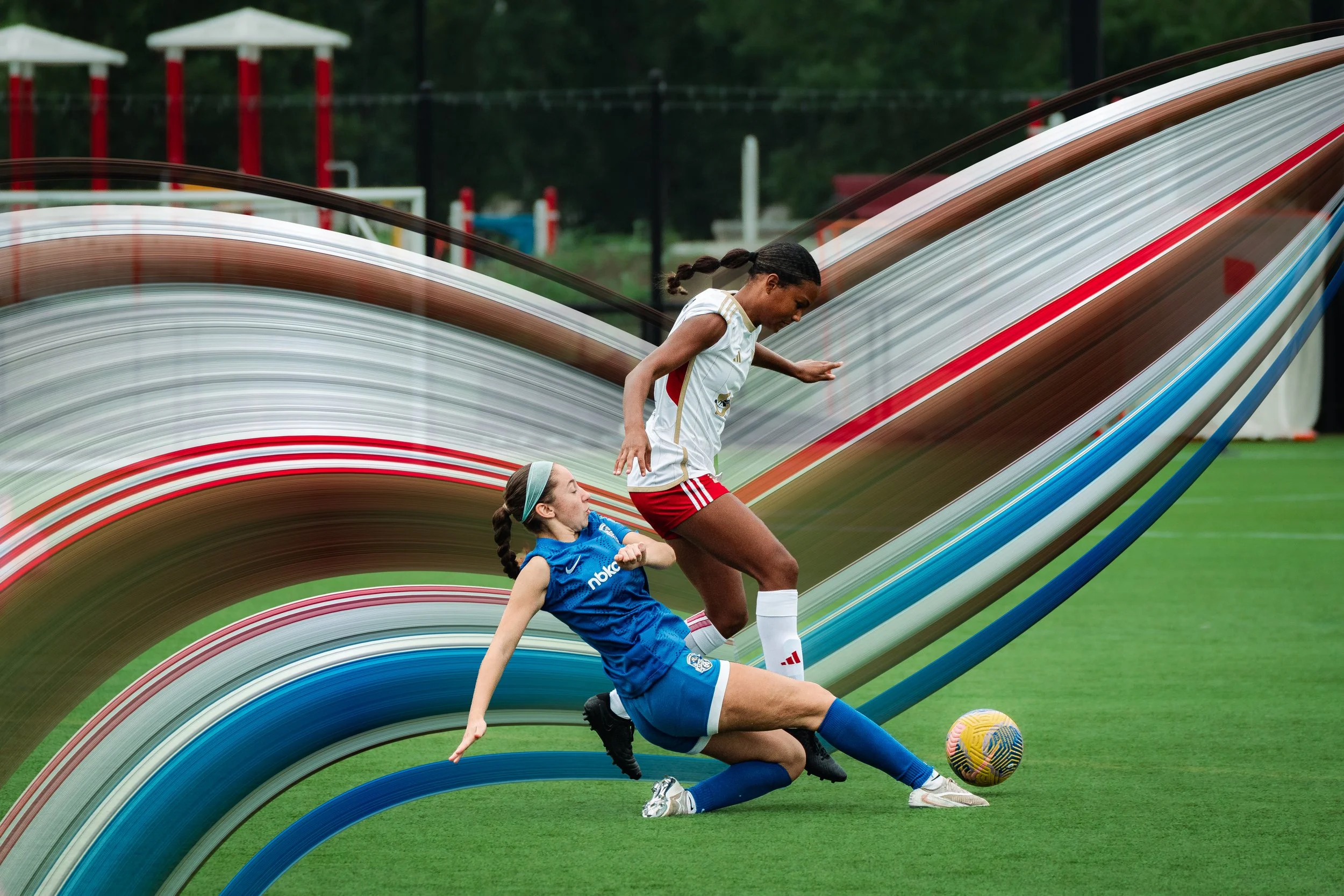 A female soccer player in a white jersey and red shorts trying to kick a yellow soccer ball while a young girl in a blue soccer uniform is falling on the grass field during a game or practice, with a background of sports field and playground equipmen