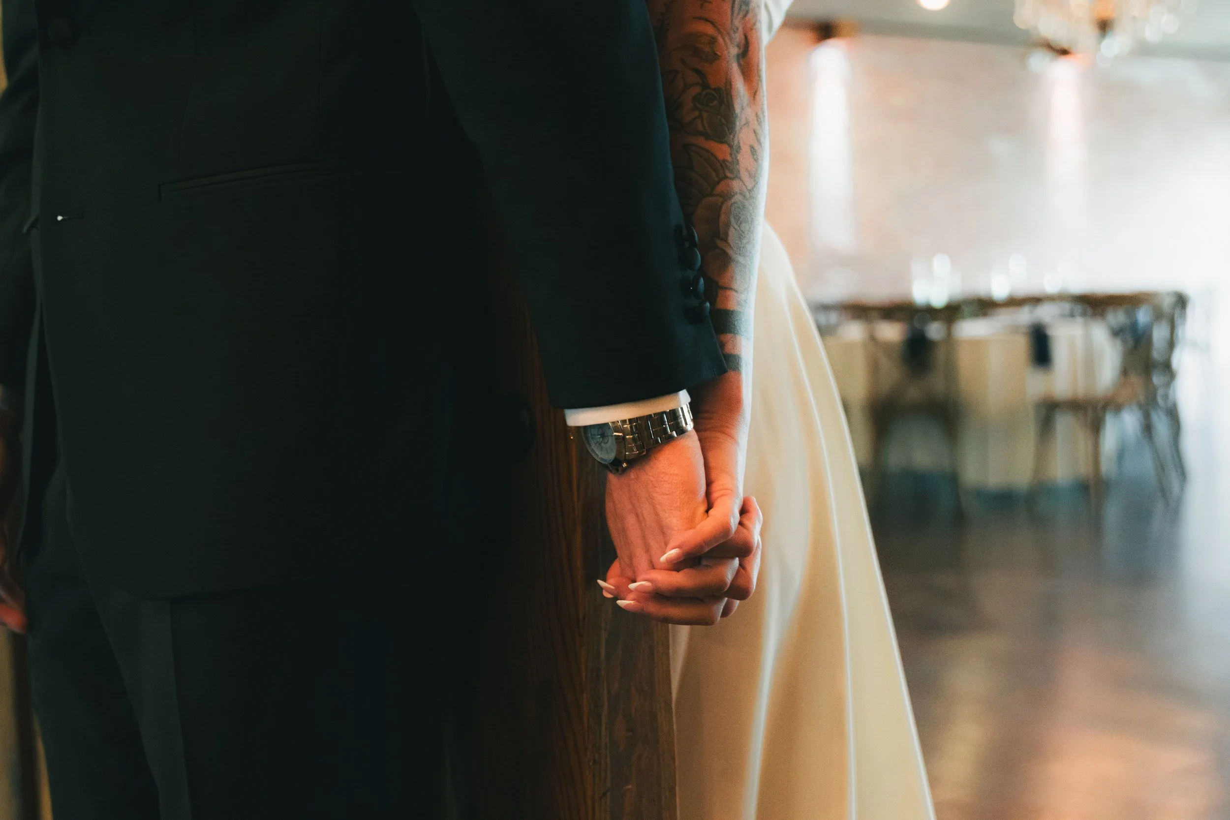Close-up of a man and a woman holding hands, with focus on their intertwined fingers. The man is wearing a dark suit and a wristwatch, and the woman is in a light-colored dress with manicured nails. The background appears to be a softly lit indoor ev