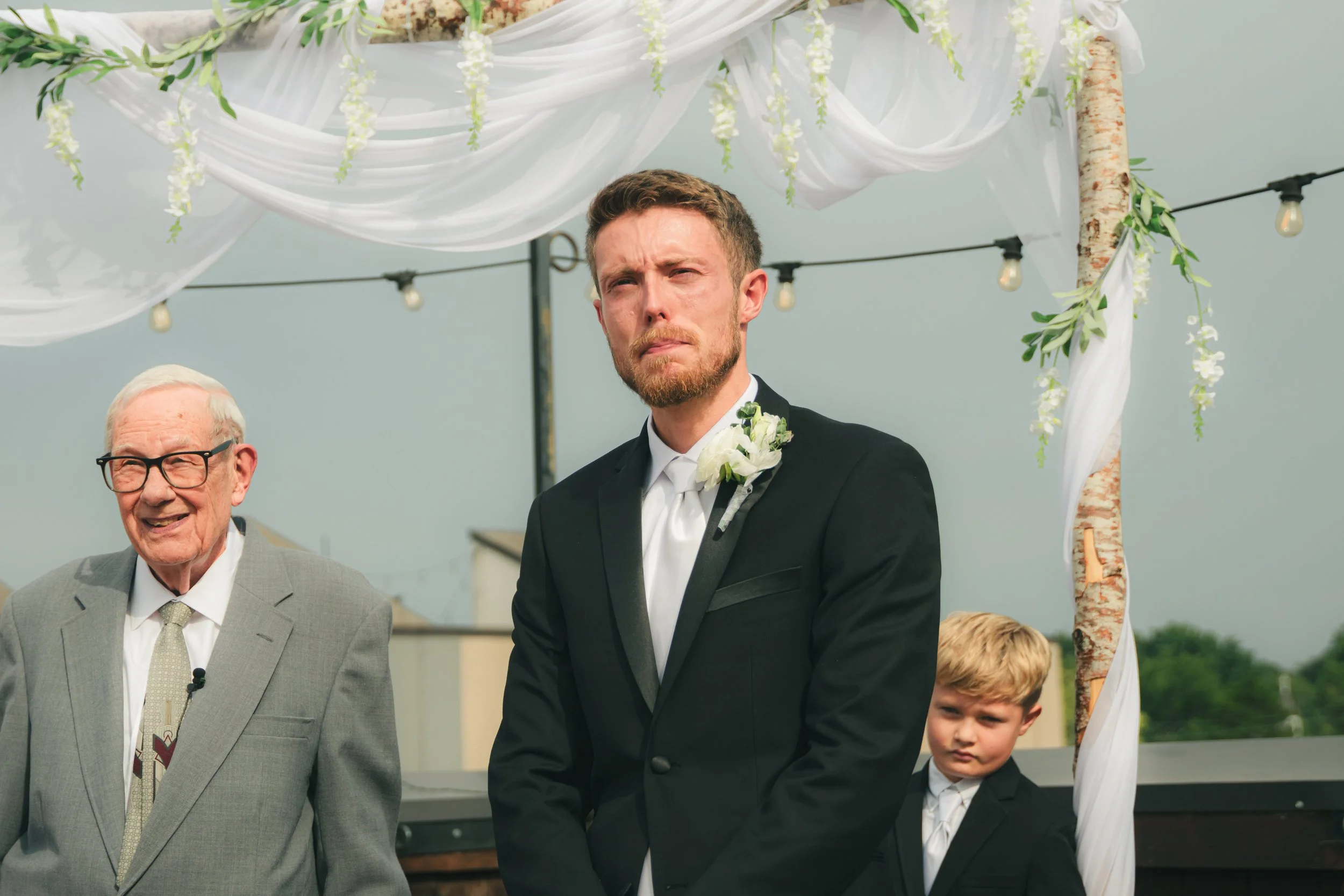 A groom in a black tuxedo with a white shirt and boutonniere stands outdoors at a wedding ceremony. An older man in a gray suit and glasses smiles nearby. Part of a child in formal attire is visible. The background features a decorated arch with whit
