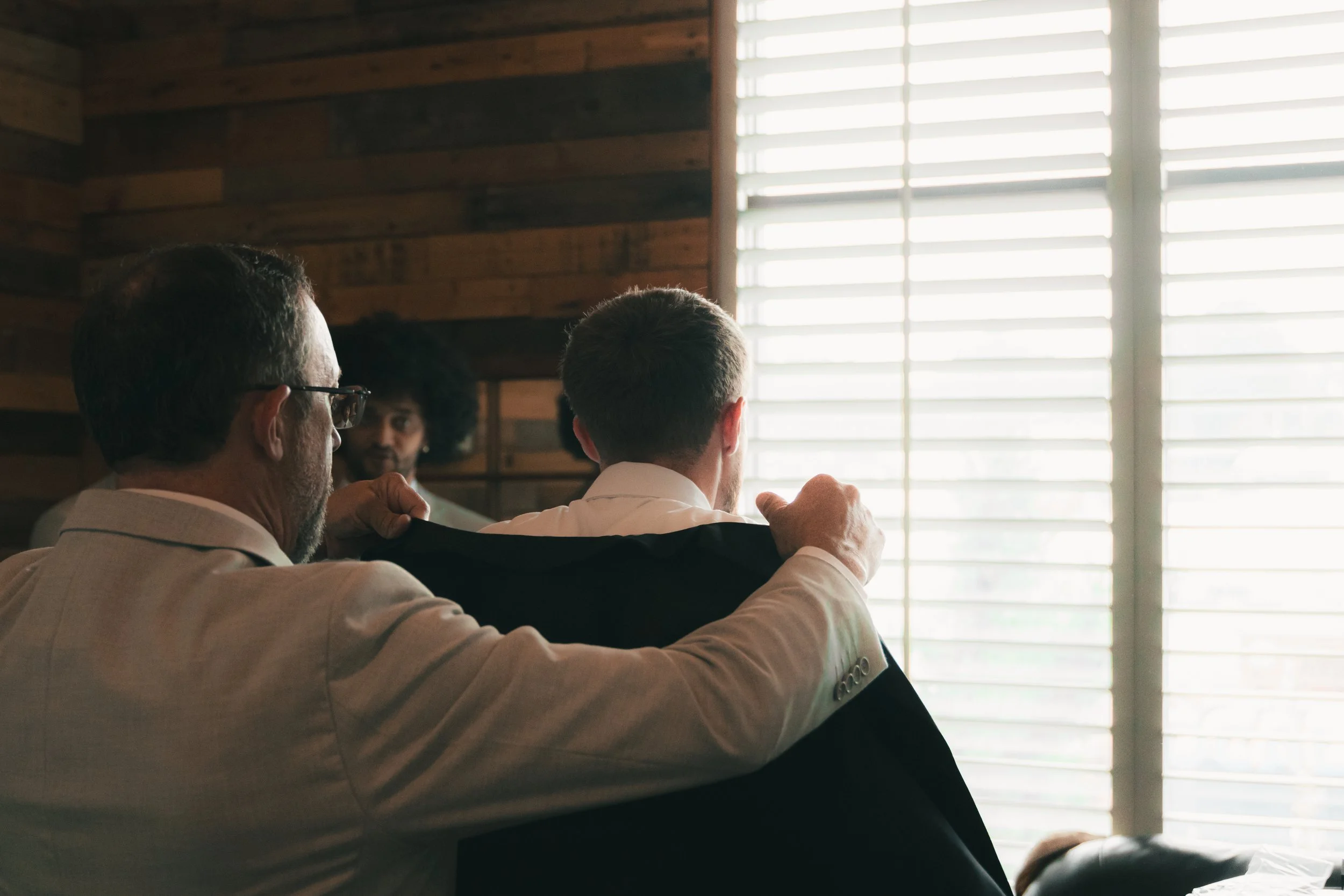 Three men in business attire sitting and talking in a room with slatted window blinds and wooden panel walls.