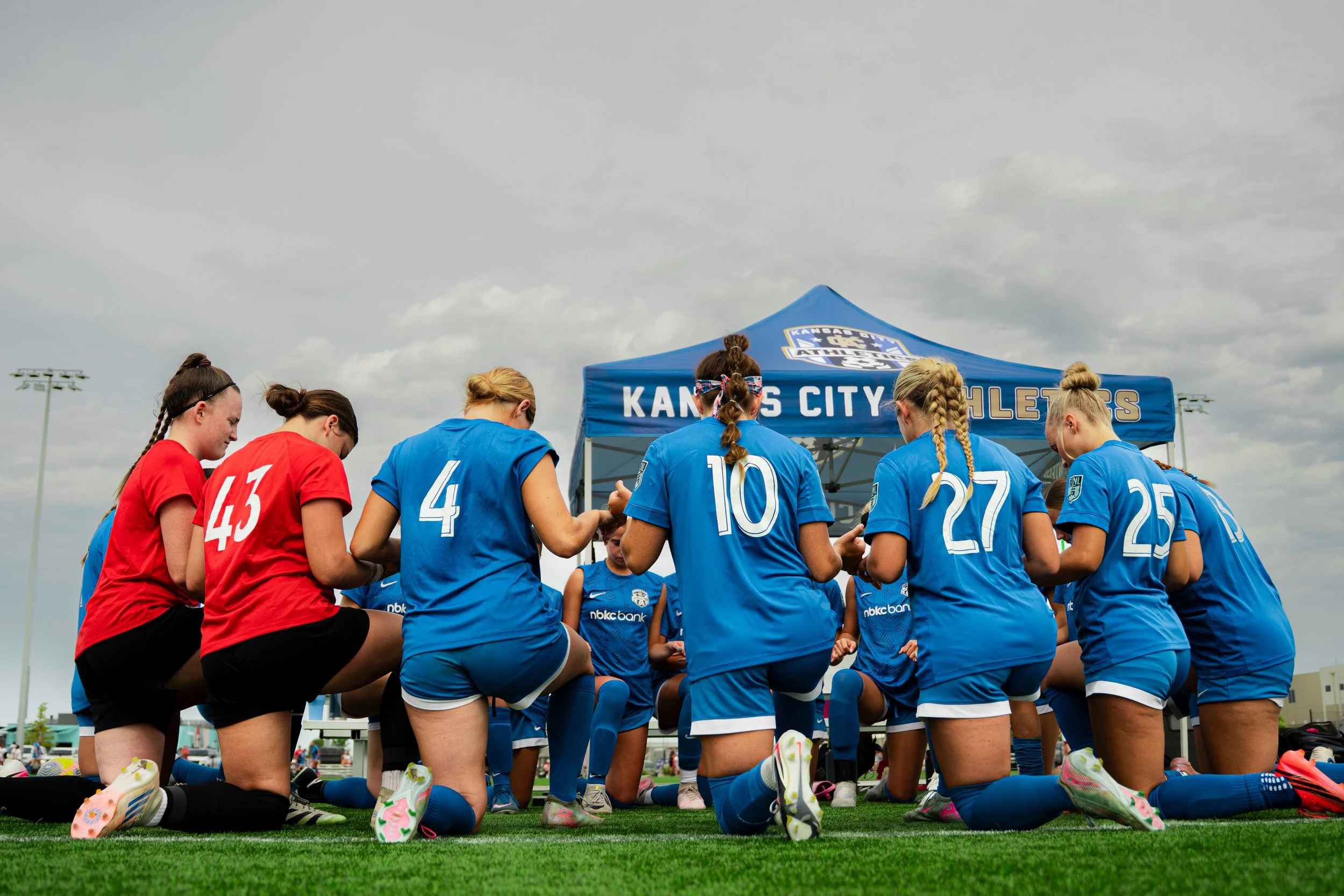 Female soccer players kneeling together in prayer, wearing red and blue uniforms, with a blue Kansas City Athletic banner in the background on a cloudy day.
