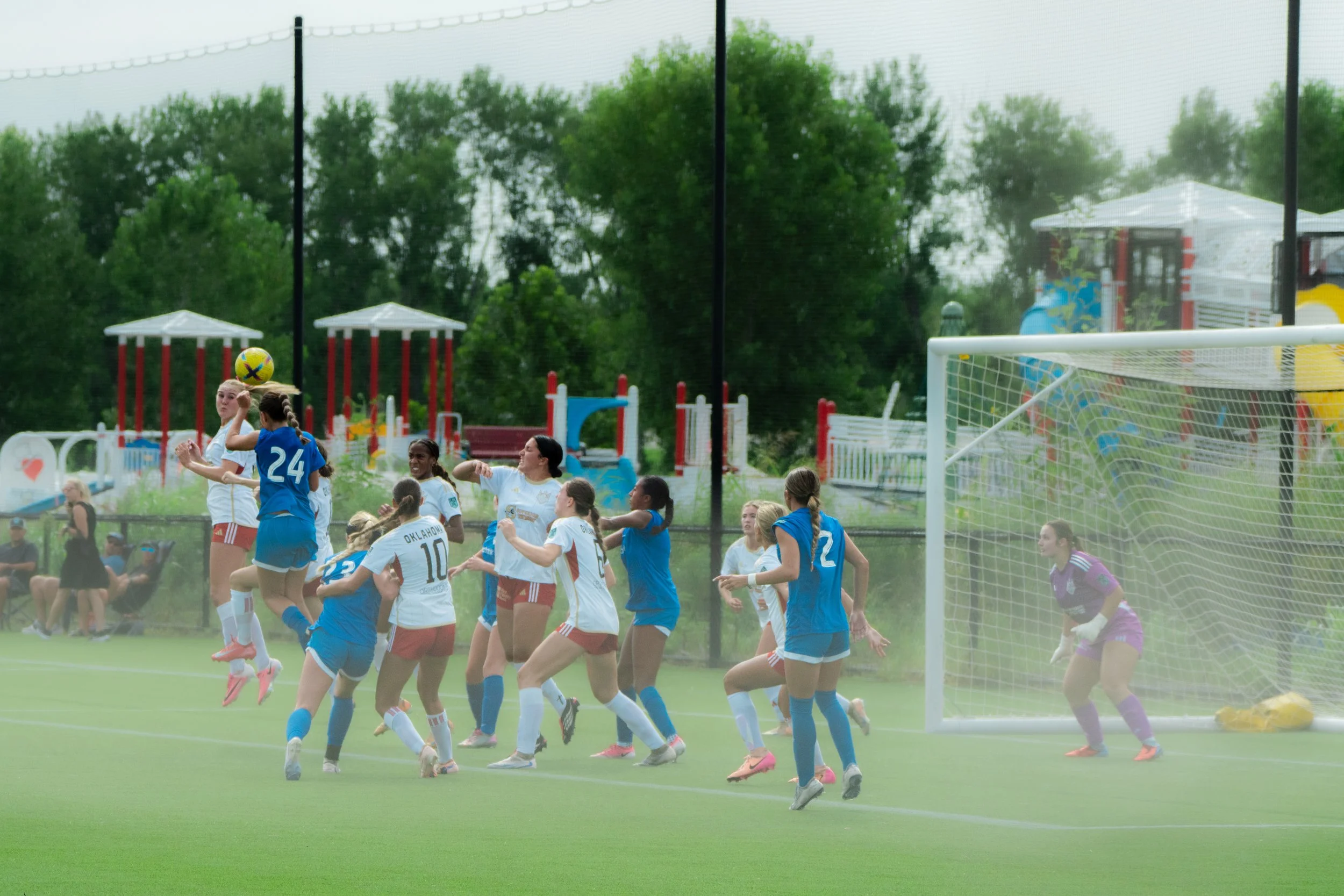 A group of women playing soccer on a field during a game, with some women wearing white jerseys and others wearing blue jerseys. One woman is in the air, heading a yellow soccer ball, near the goalpost with a goalkeeper in purple attempting to block 