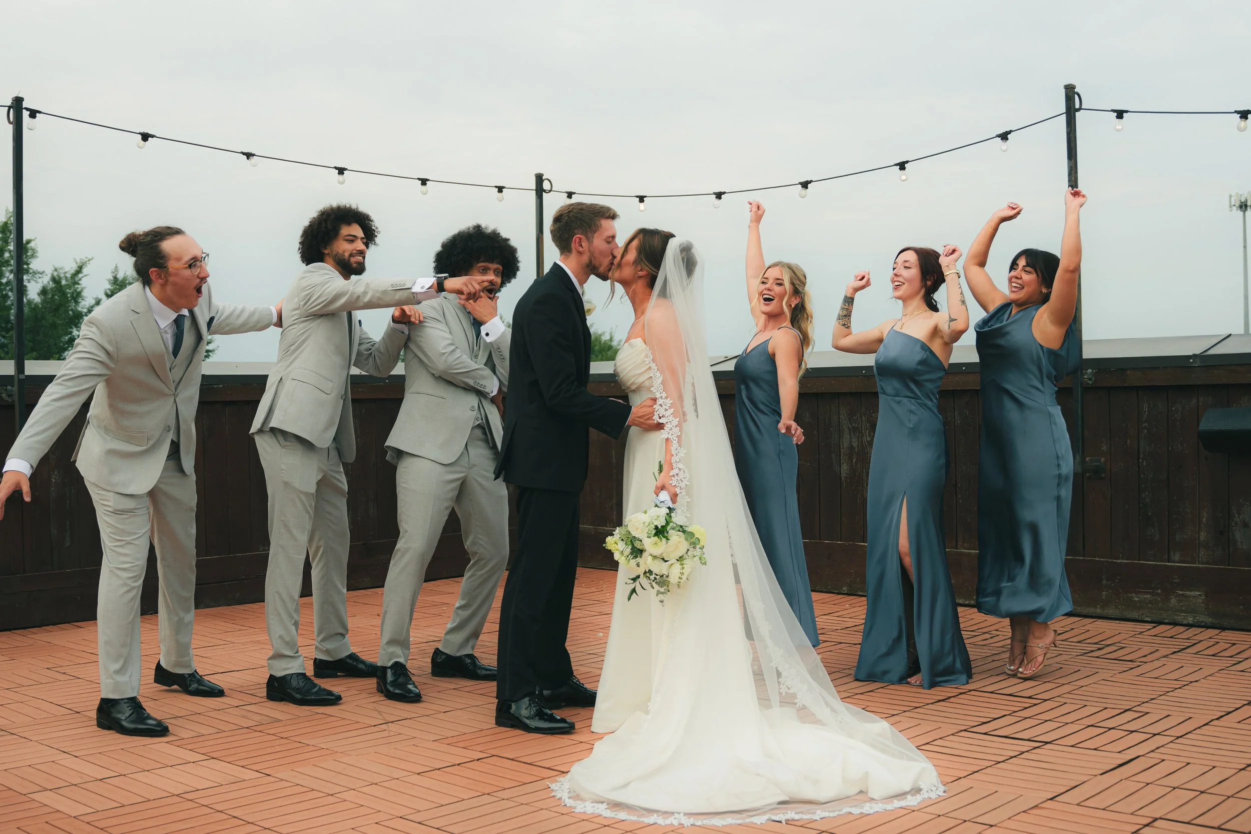 A wedding ceremony with a bride and groom kissing, surrounded by six bridesmaids and groomsmen on a rooftop patio with string lights, celebrating with raised fists and smiles.