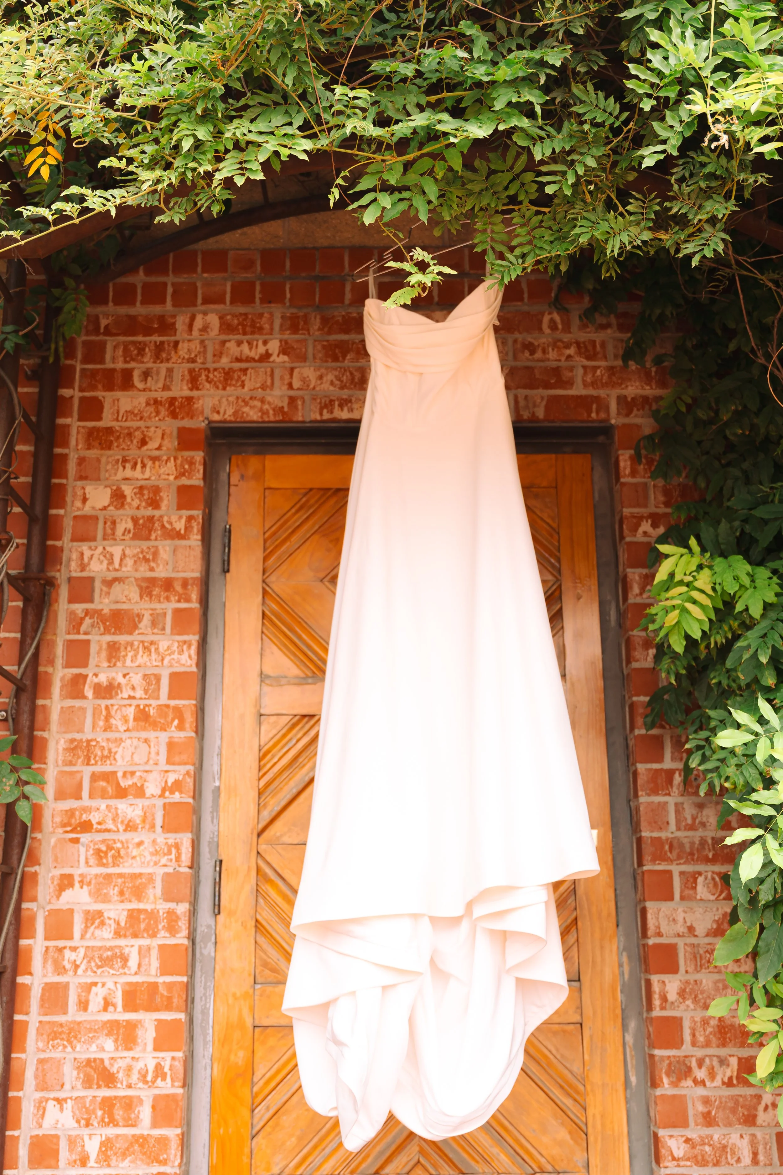 A white wedding dress hanging from a vine-covered archway in front of a wooden door and brick wall.