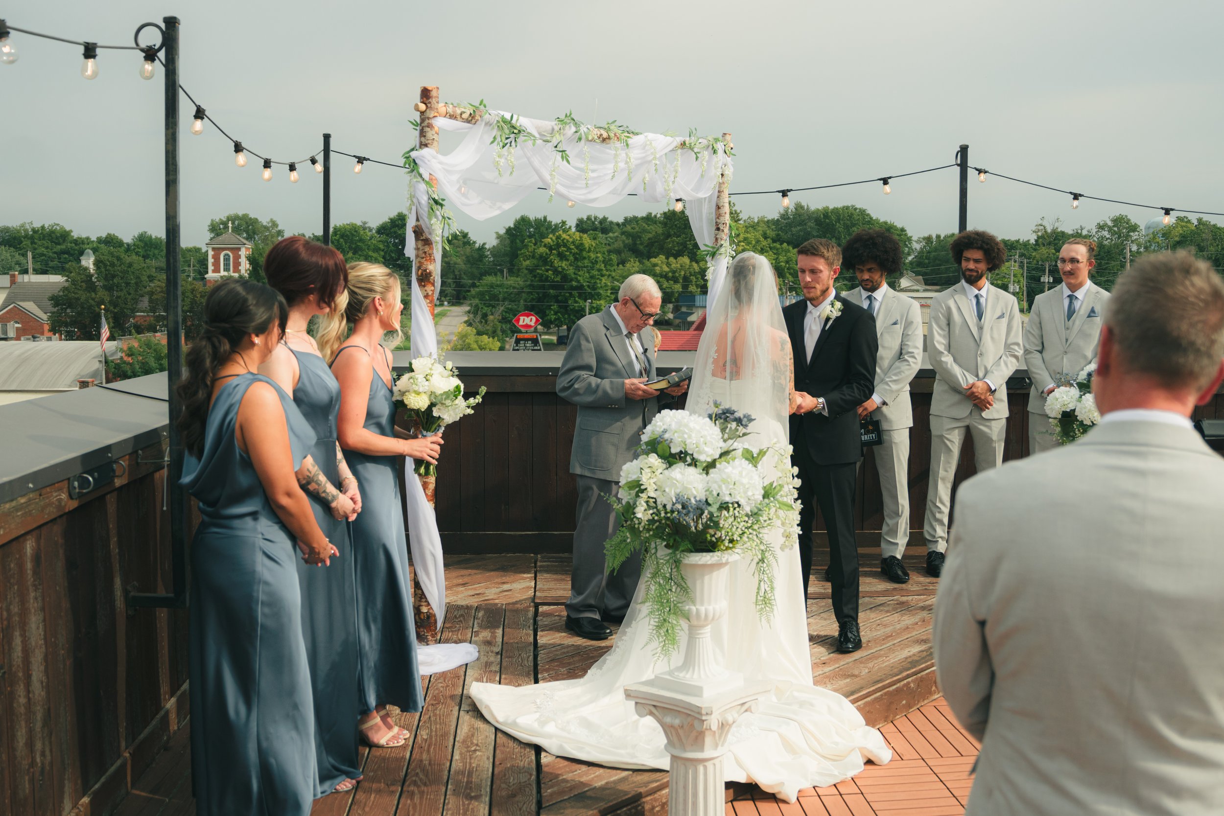 A wedding ceremony taking place outdoors on a rooftop, with a bride and groom standing in front of an officiant. Bridesmaids and groomsmen are standing beside them, and the setting is decorated with string lights, white drapery, and floral arrangemen