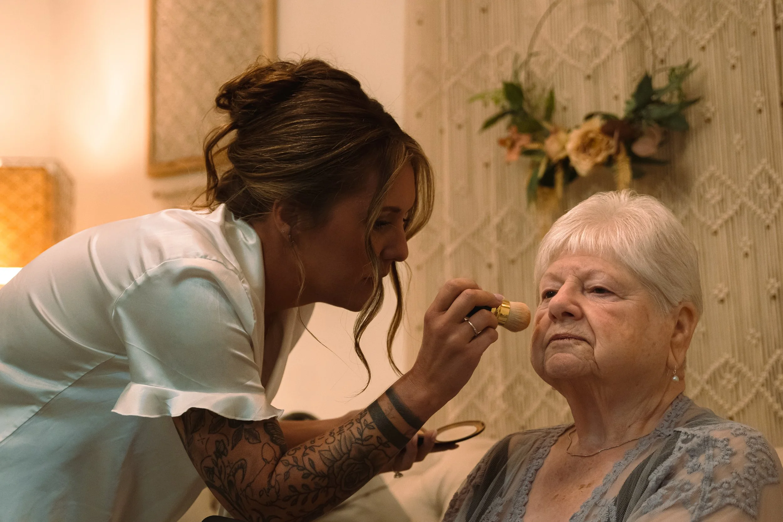 A makeup artist applying makeup to an elderly woman with white hair in a room with warm lighting and floral decorations.