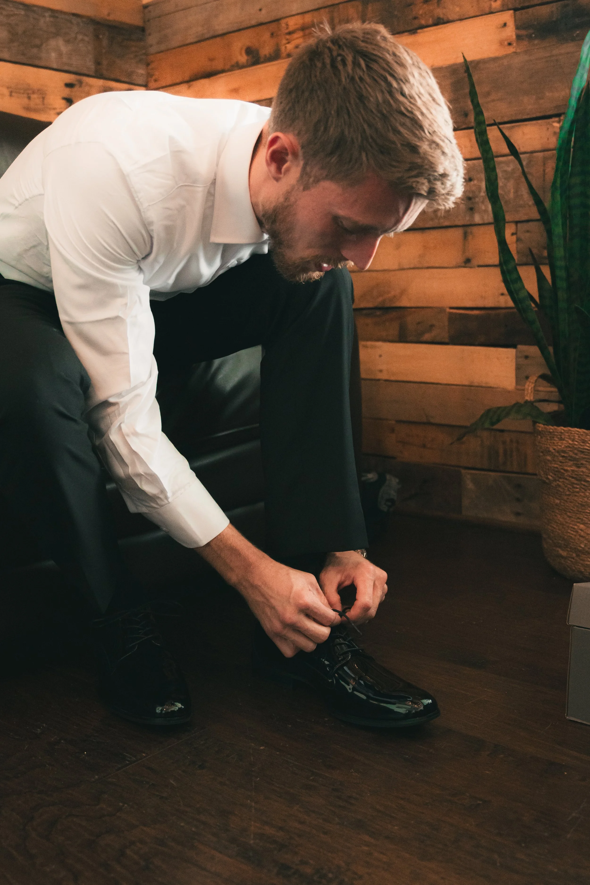 A man dressed in a white shirt and black pants is sitting on a chair, leaning forward, as he puts on or adjusts his black dress shoes. He is in a room with wooden walls and a tall green plant nearby.