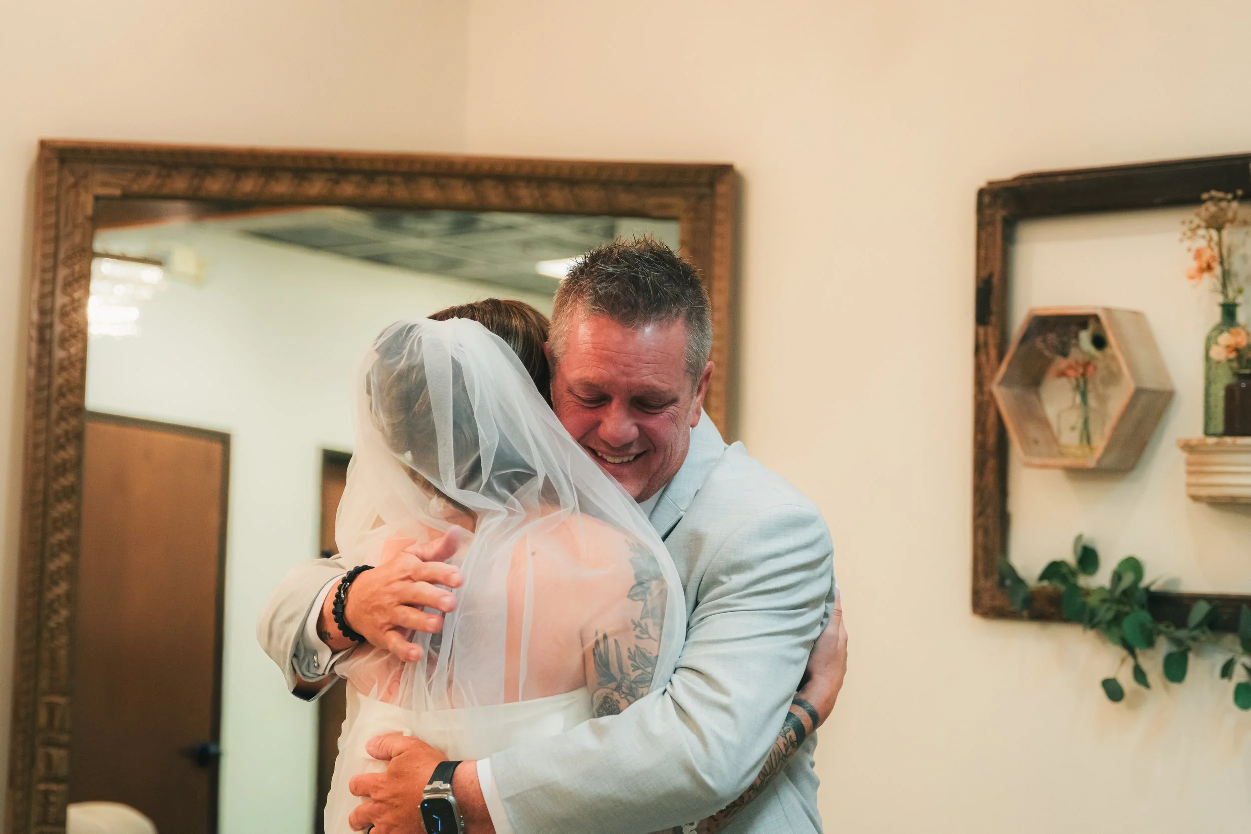 A man and woman hugging each other, smiling, at a celebration or wedding in a decorated room with mirrors and framed artwork.
