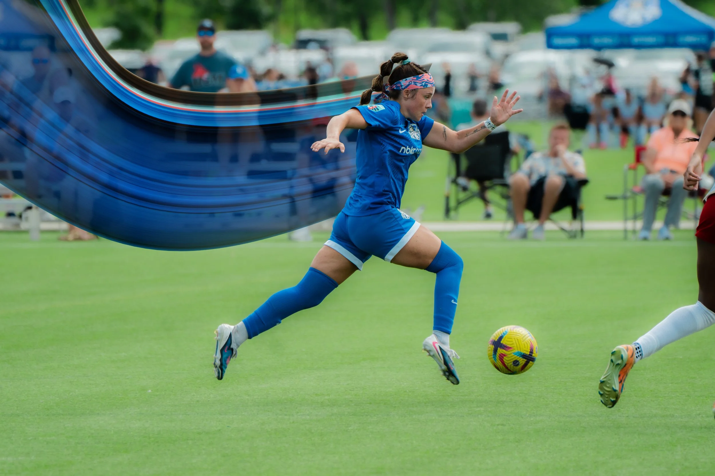 A female soccer player in a blue uniform with a colorful headband is running on a soccer field, chasing after a multicolored soccer ball during a game or practice. There are spectators and other players in the background.