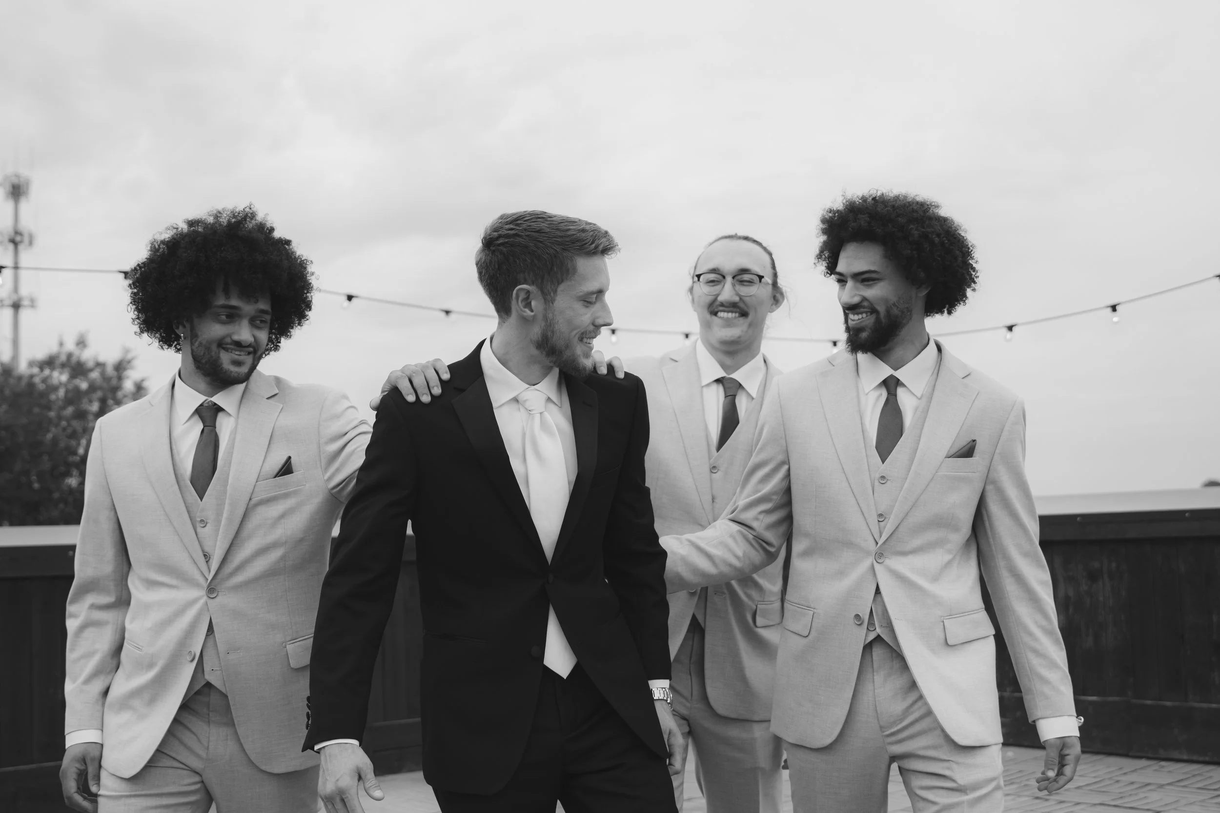 Four men in suits walking together outdoors, with the man in the center wearing a black suit and the other three in light-colored suits. They are smiling and appear to be having a friendly conversation, with one man placing his hand on another's shou