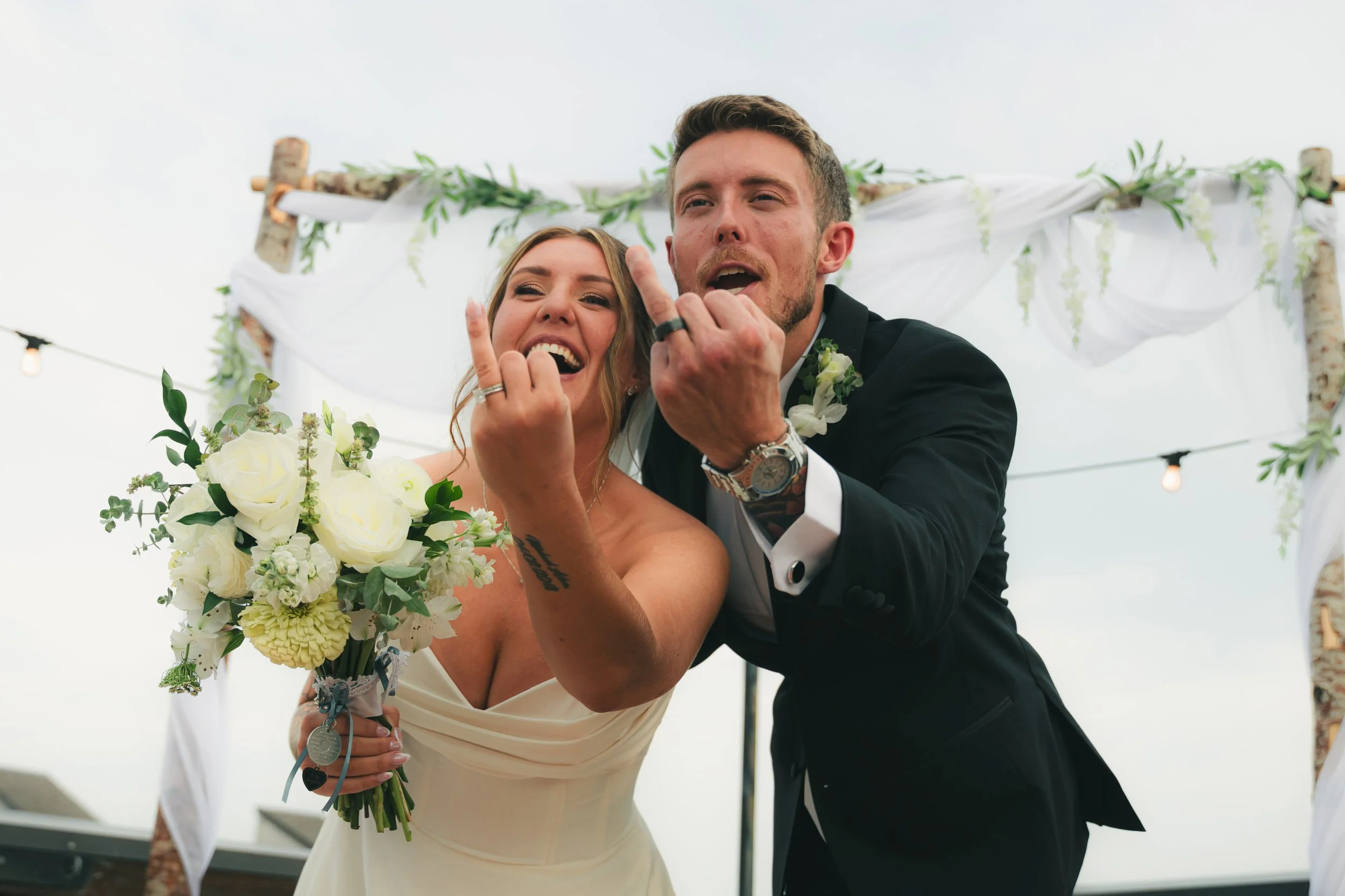 A bride and groom celebrating at their wedding, smiling and making hand gestures while holding a bouquet of white flowers, under a decorated outdoor wedding arch.